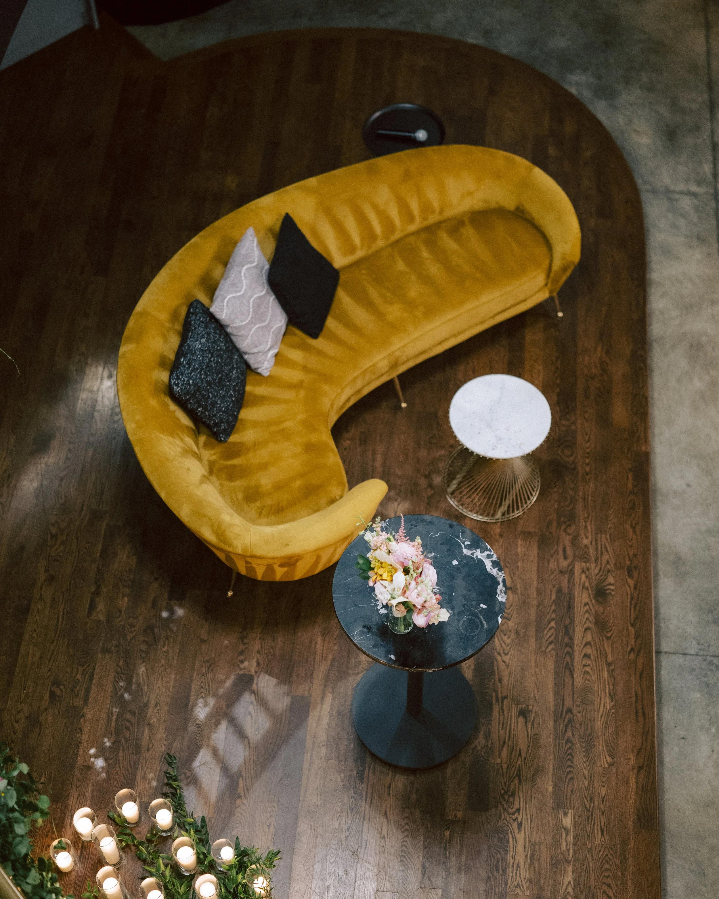 A yellow velvet curved sofa with three decorative pillows, a small round marble-top side table, and a black marble coffee table with a flower arrangement on it. There are candles on the floor near the sofa.