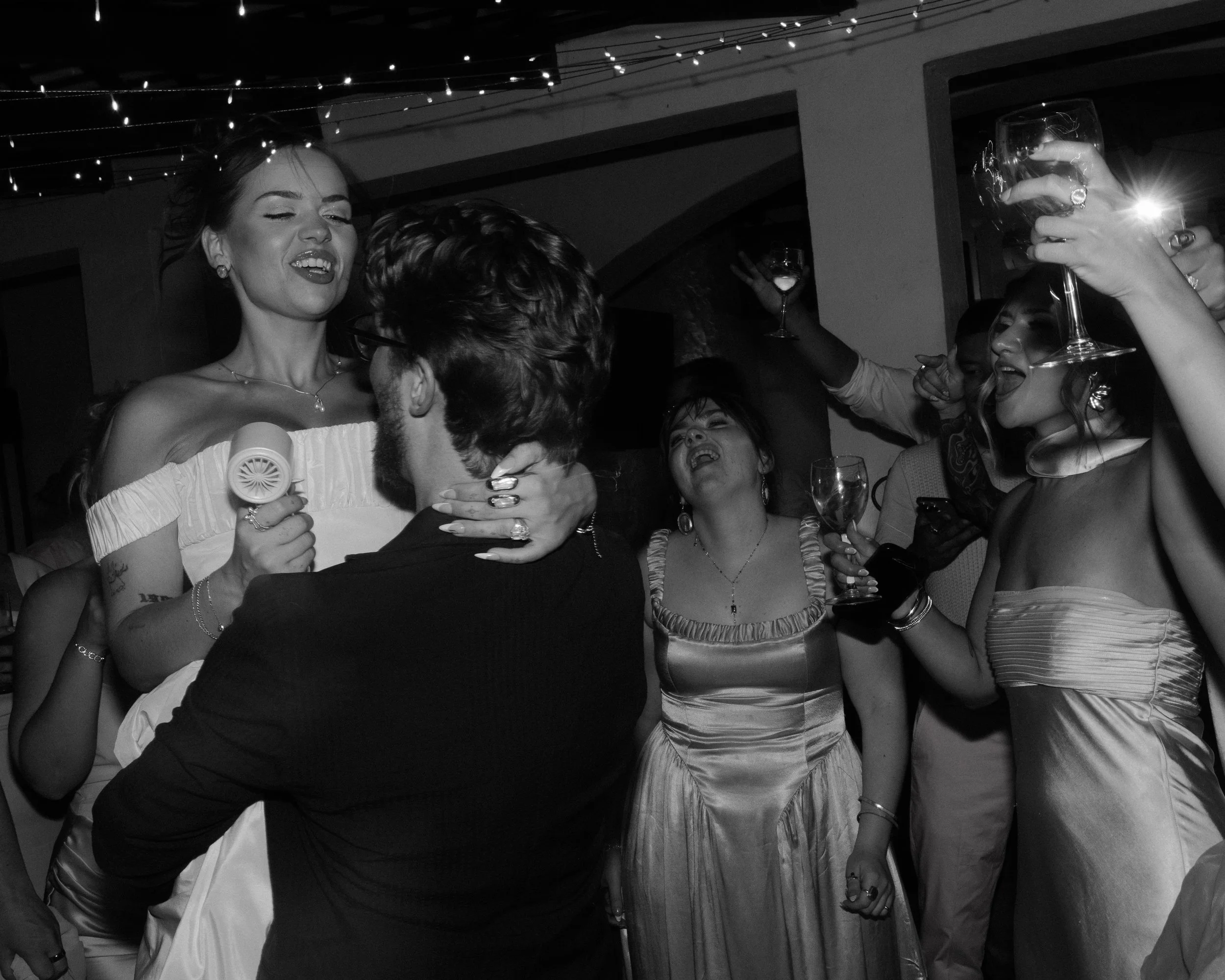 People celebrating at a wedding reception, dancing and holding glasses of wine in a festive atmosphere, black and white photograph.