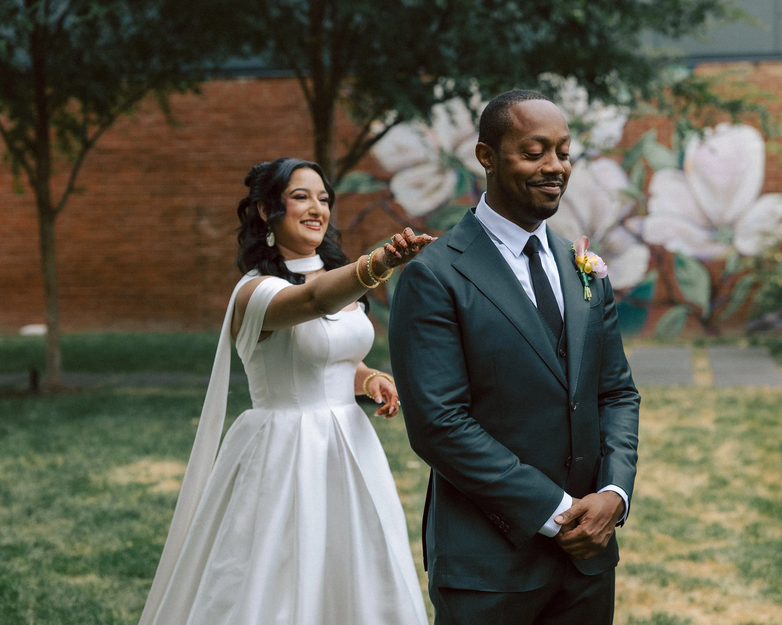 A bride with dark hair styled in loose curls is adjusting the groom's tie. She is wearing a white wedding dress with sleeves and jewelry. The groom, with a boutonniere on his suit, is standing in front of her with his eyes closed, smiling softly. The
