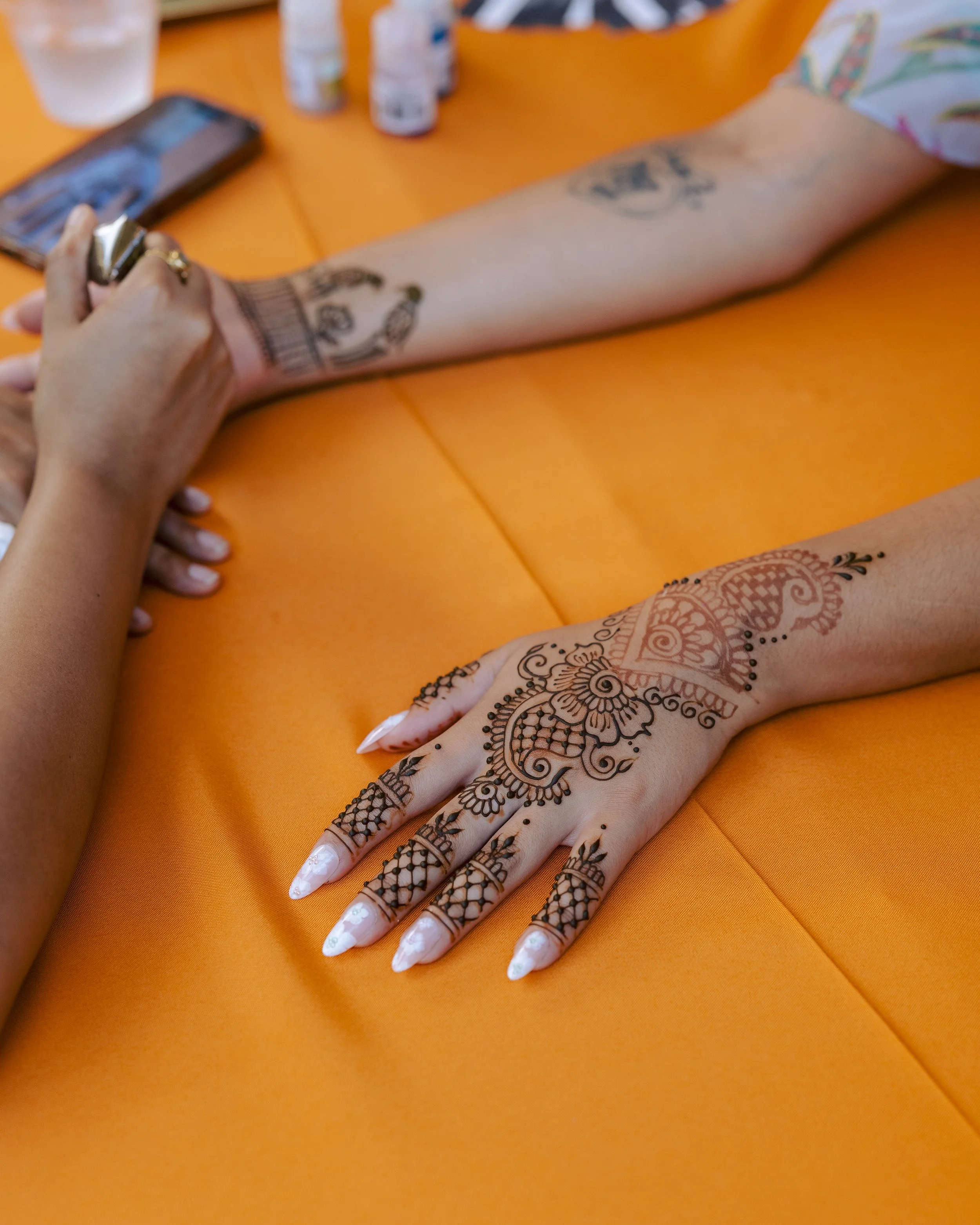 A person getting a henna tattoo on their hand and forearm at a colorful event, with a bright orange tablecloth in the background.