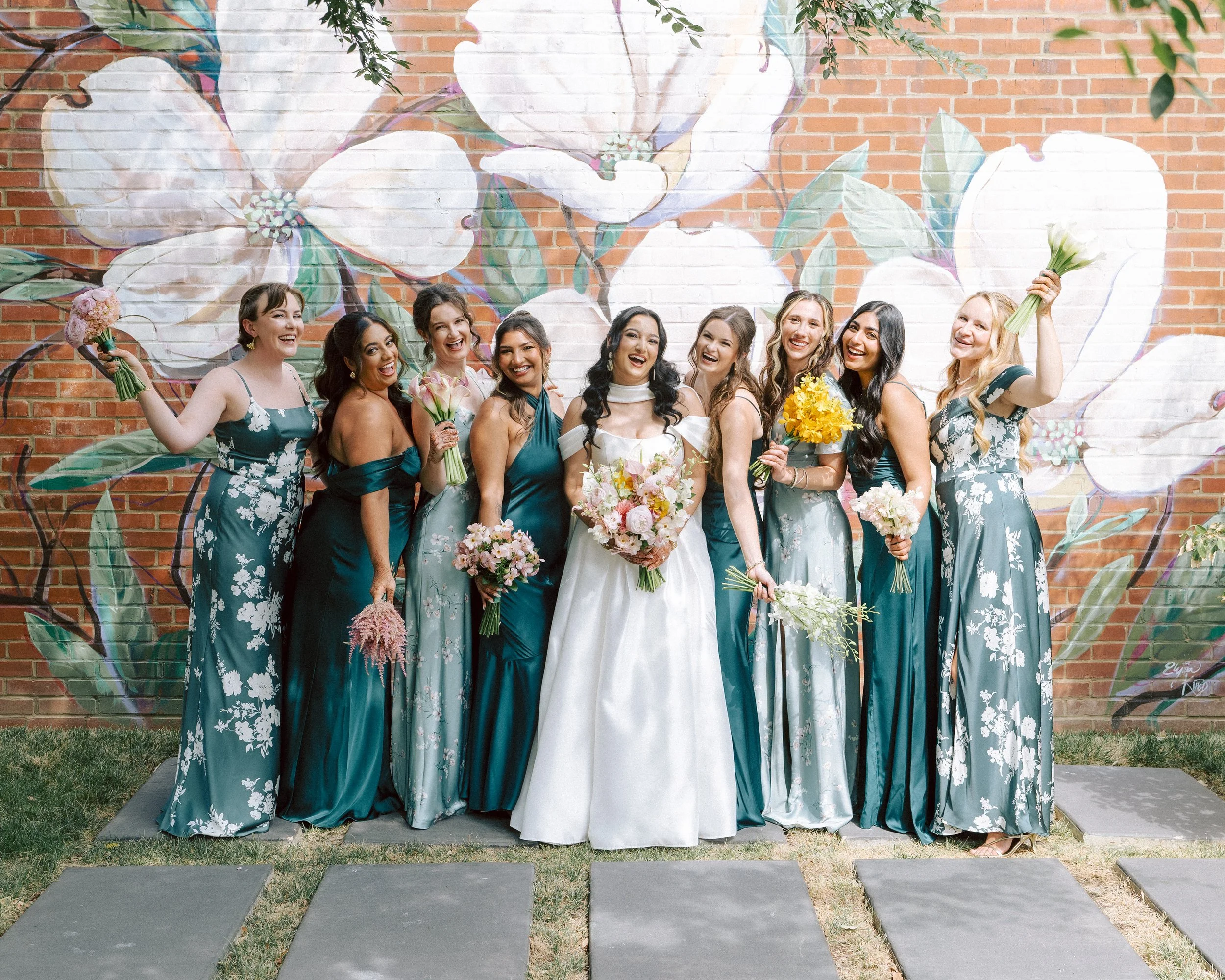 A group of women, including a bride in a white wedding dress, standing in front of a colorful floral mural on a brick wall, holding bouquets of flowers and smiling.