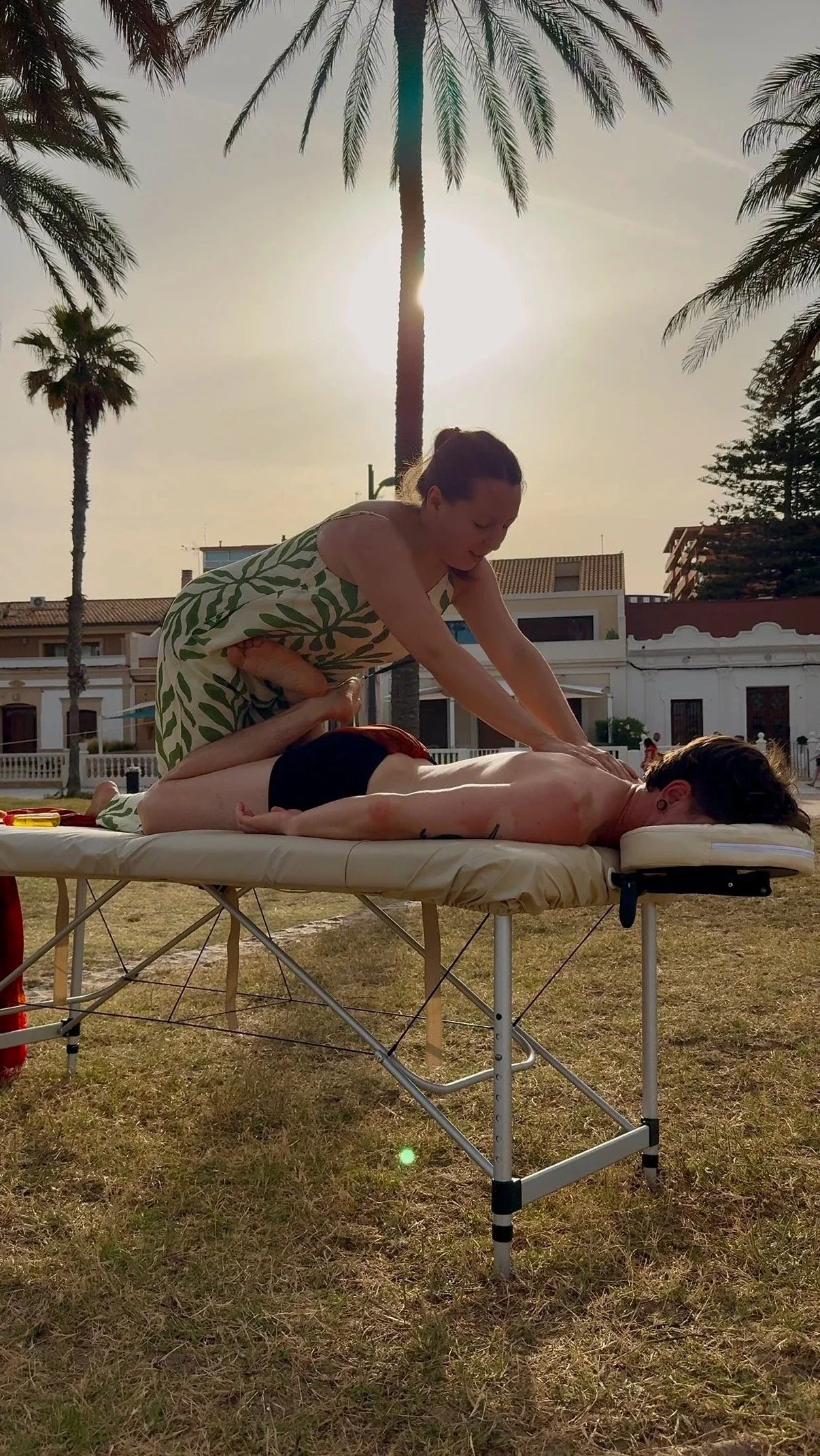 A woman receives a massage outdoors on a massage table with palm trees and buildings in the background, during sunset.
