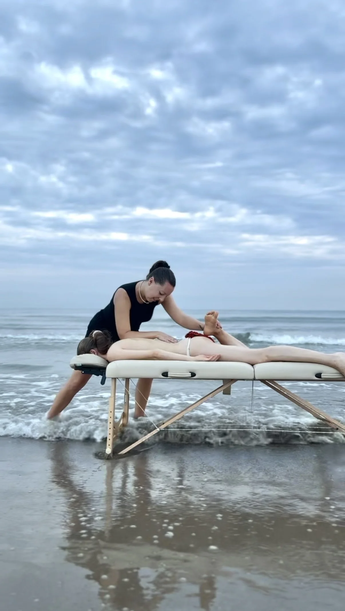 A massage therapist giving a  massage on the Cabanyal beach in Valencia, with sea waves and cloudy sky in the background.