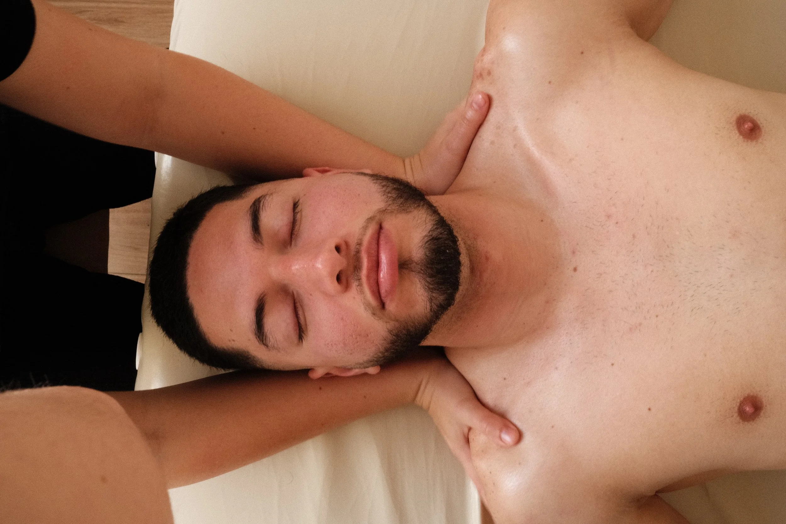 A man with short black hair and a beard receiving a massage with his eyes closed, being gently touched on his shoulders and neck by a person with lightly tanned skin.