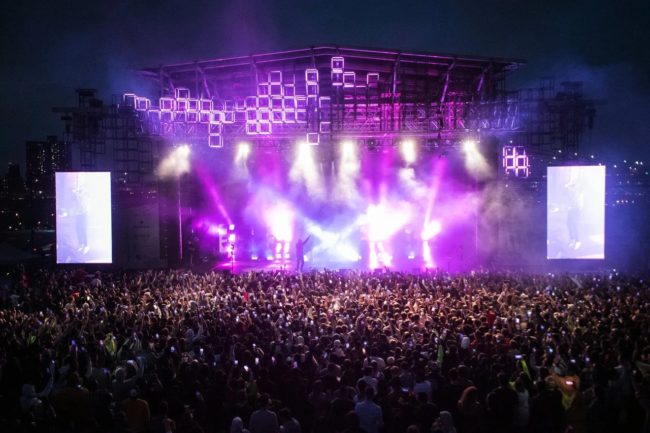 Nighttime outdoor concert with a large crowd, bright purple stage lighting, and two large screens on either side of the stage.