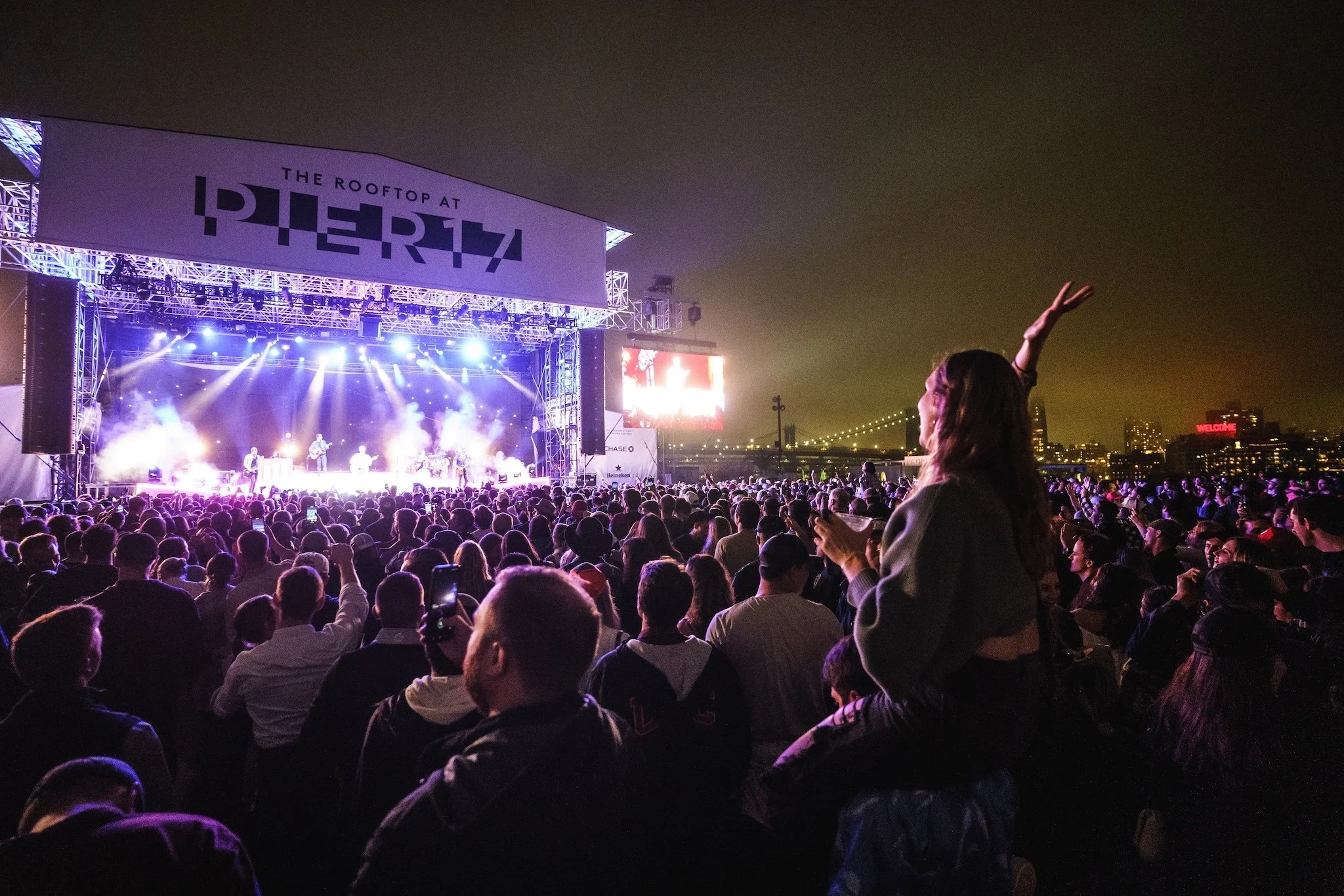 Crowd attending outdoor music concert at night with stage, lights, and city skyline in background