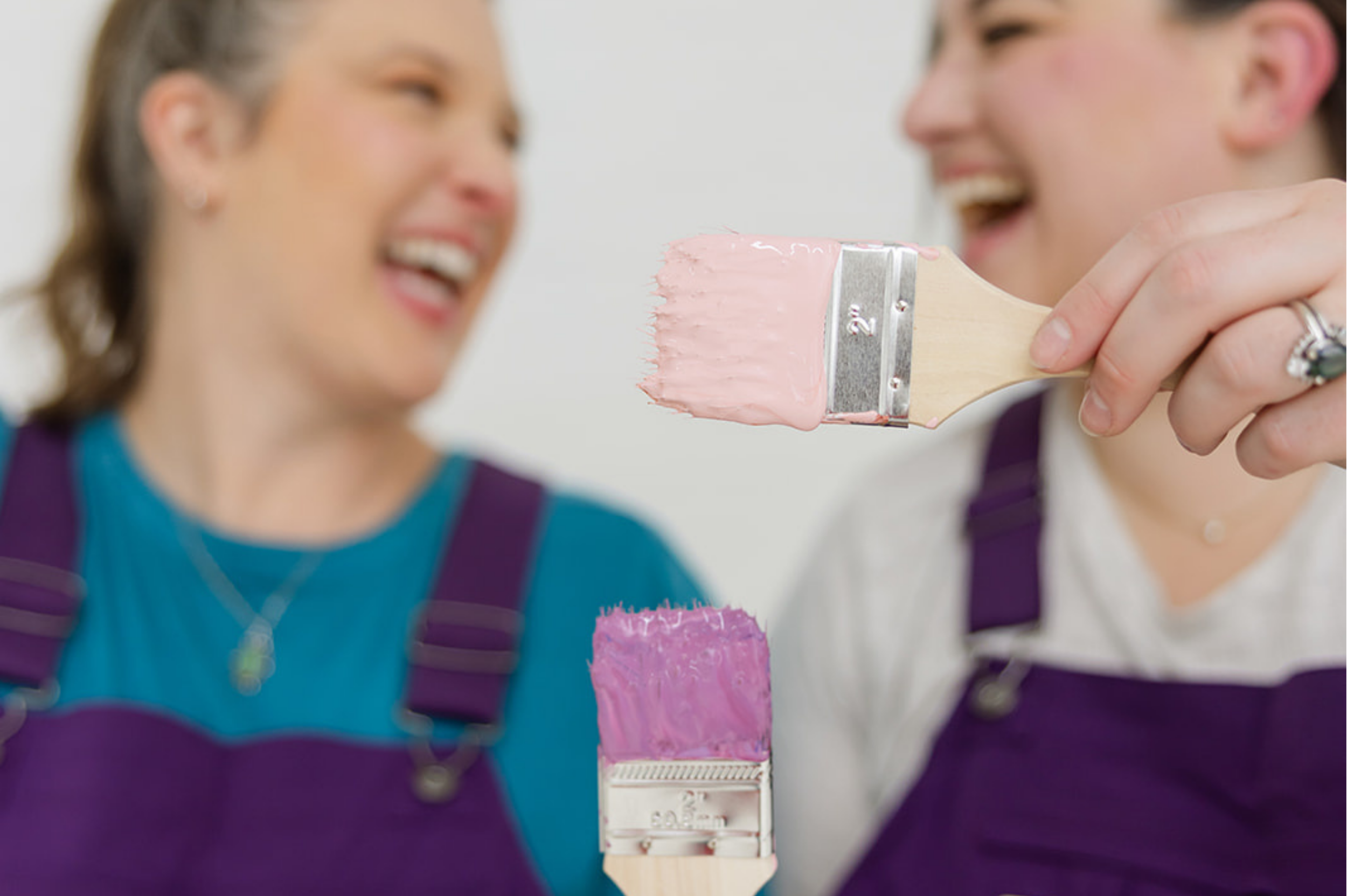 Founders of The List in purple overalls holding paintbrushes with pink and purple paint, showcasing the playful and professional brand personality designed by Allie Marie Design.