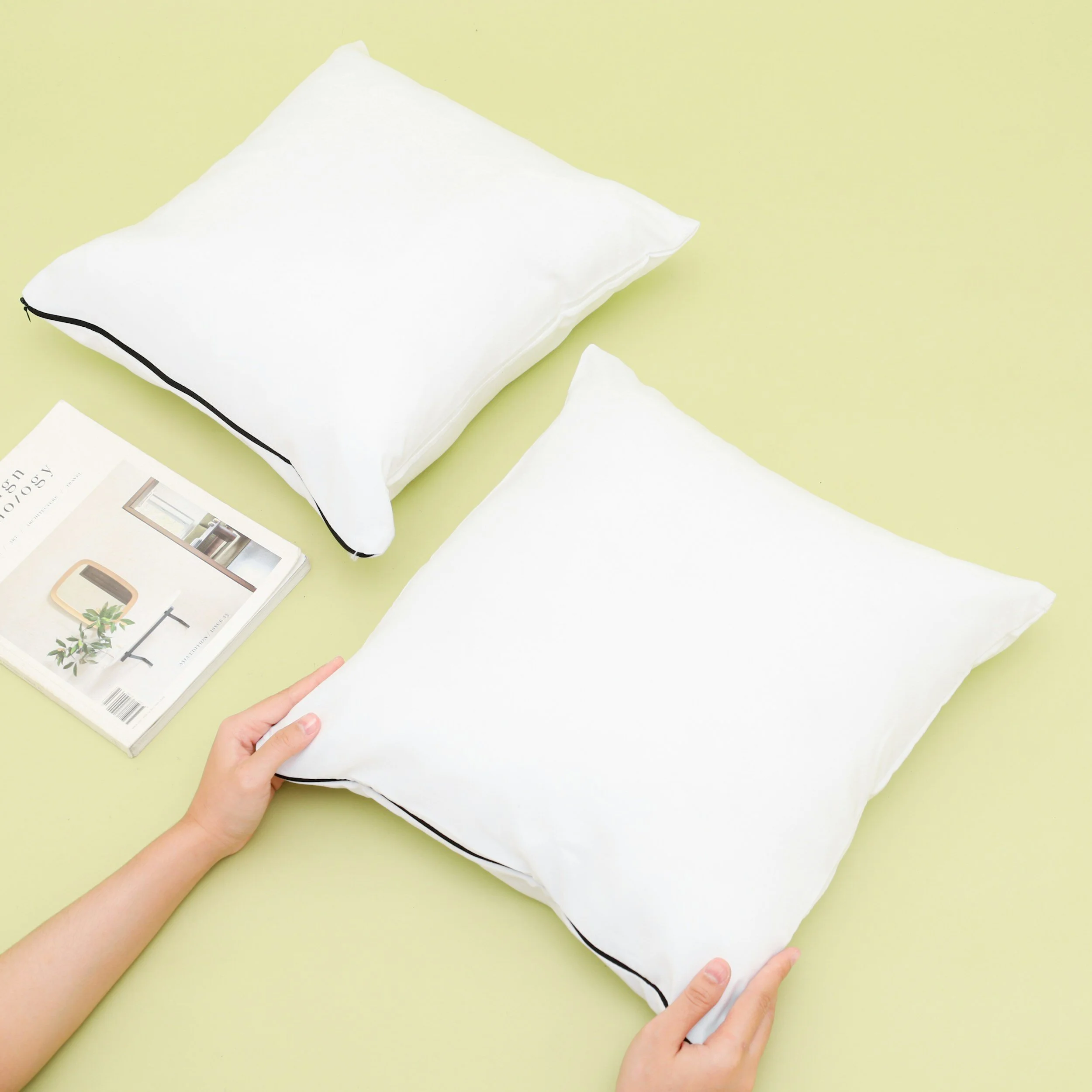 Two white pillows with black piping on a light green surface. A person is holding one pillow. A magazine is placed nearby.