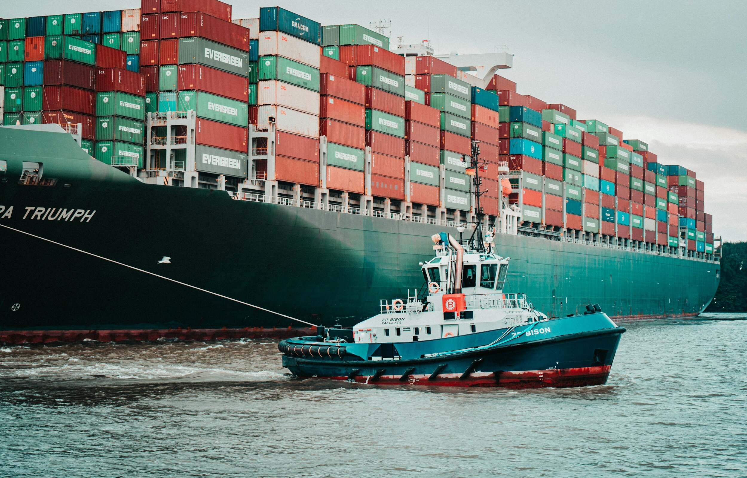 A large cargo ship with multiple stacked shipping containers sailing in water, accompanied by a small tugboat named BISON in front.
