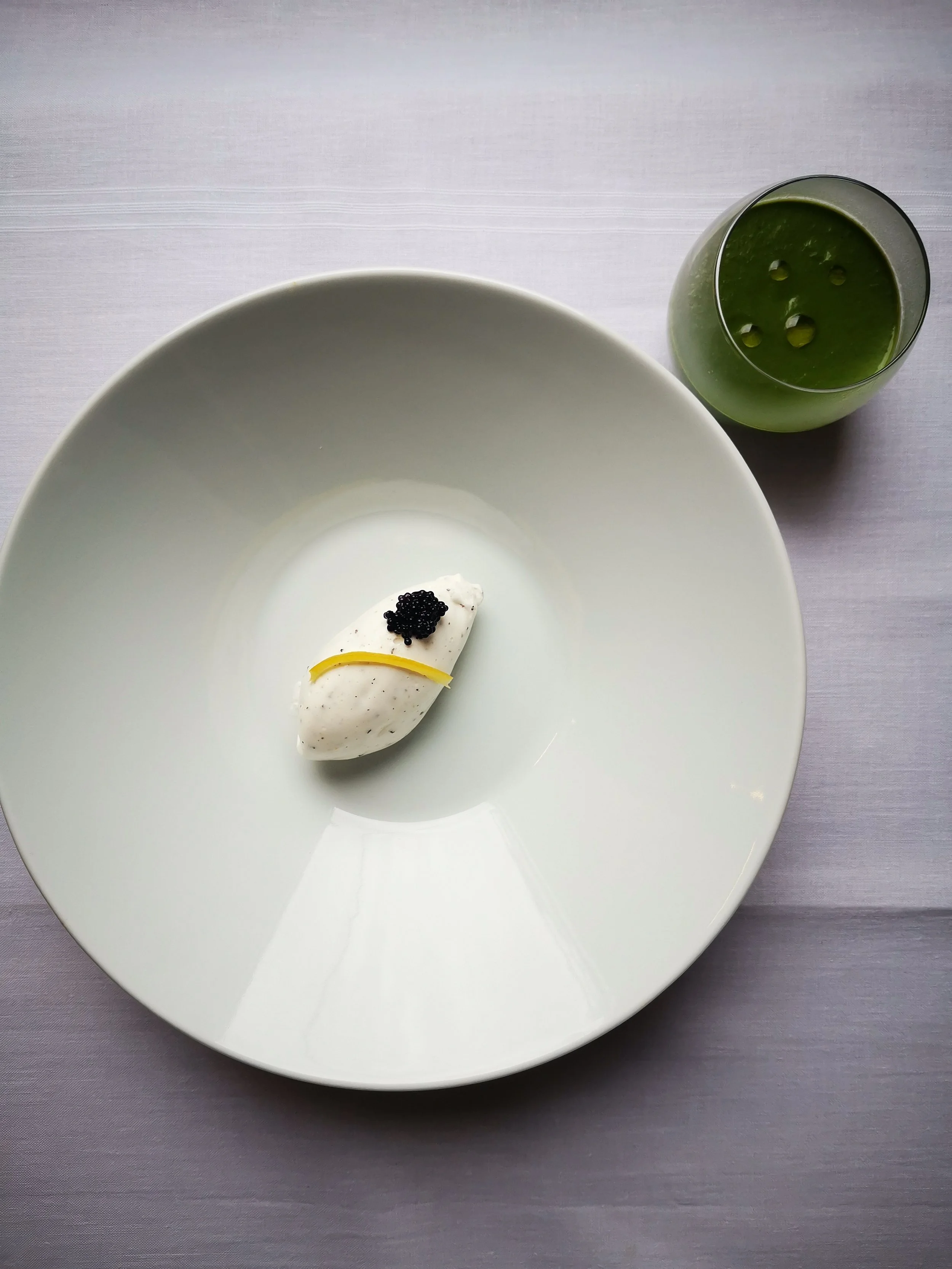 Un plat minimaliste avec une quenelle de glace garnie d'un zeste de citron et de caviar, accompagné d'un verre de liquide vert.
