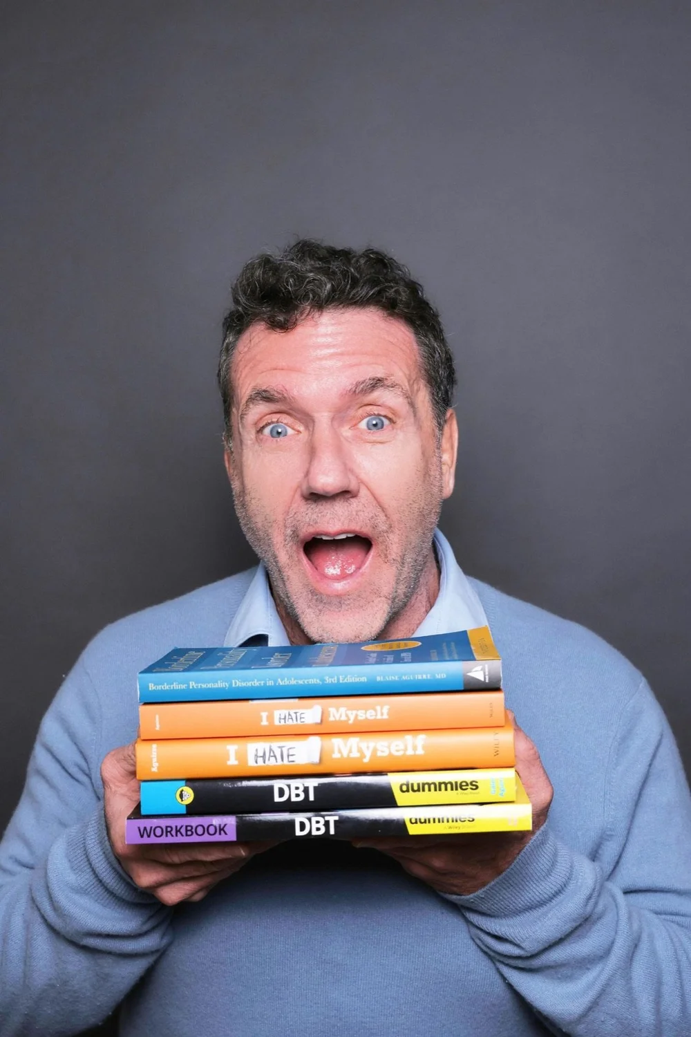 Dr. Blaise Aguirre with an exaggerated surprised expression, holding a stack of his books