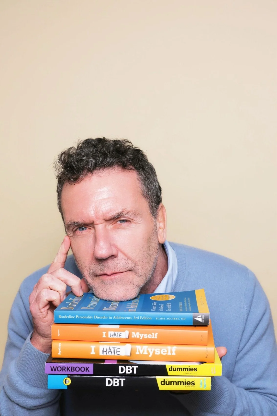 Dr. Blaise Aguirre resting his chin on a stack of his books, including I Hate Myself and DBT For Dummies