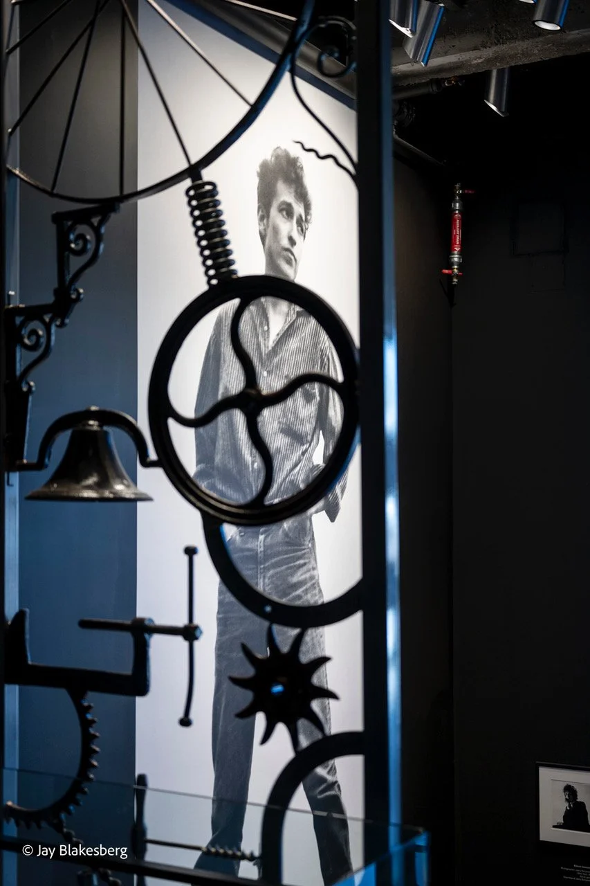 Black and white photo of a young man with styled hair, wearing a striped shirt and light-colored pants, seen through a decorative metal sculpture with gears and circular patterns, in an art exhibit setting.