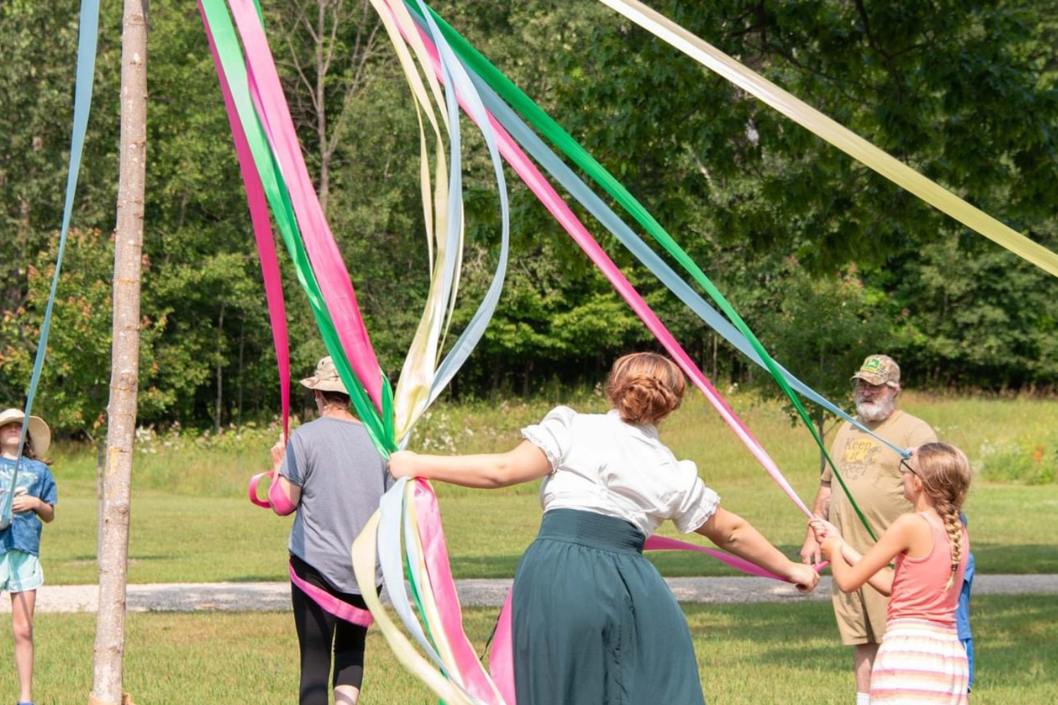 Children and adults playing with long colorful ribbons on a grassy outdoor field with trees in the background.
