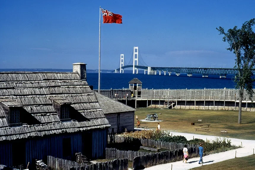 A historic fort site featuring a wooden building with a shingled roof, an American flag flying on a flagpole, a grassy area with picnic tables, and a view of the Mackinac bridge over water in the background.