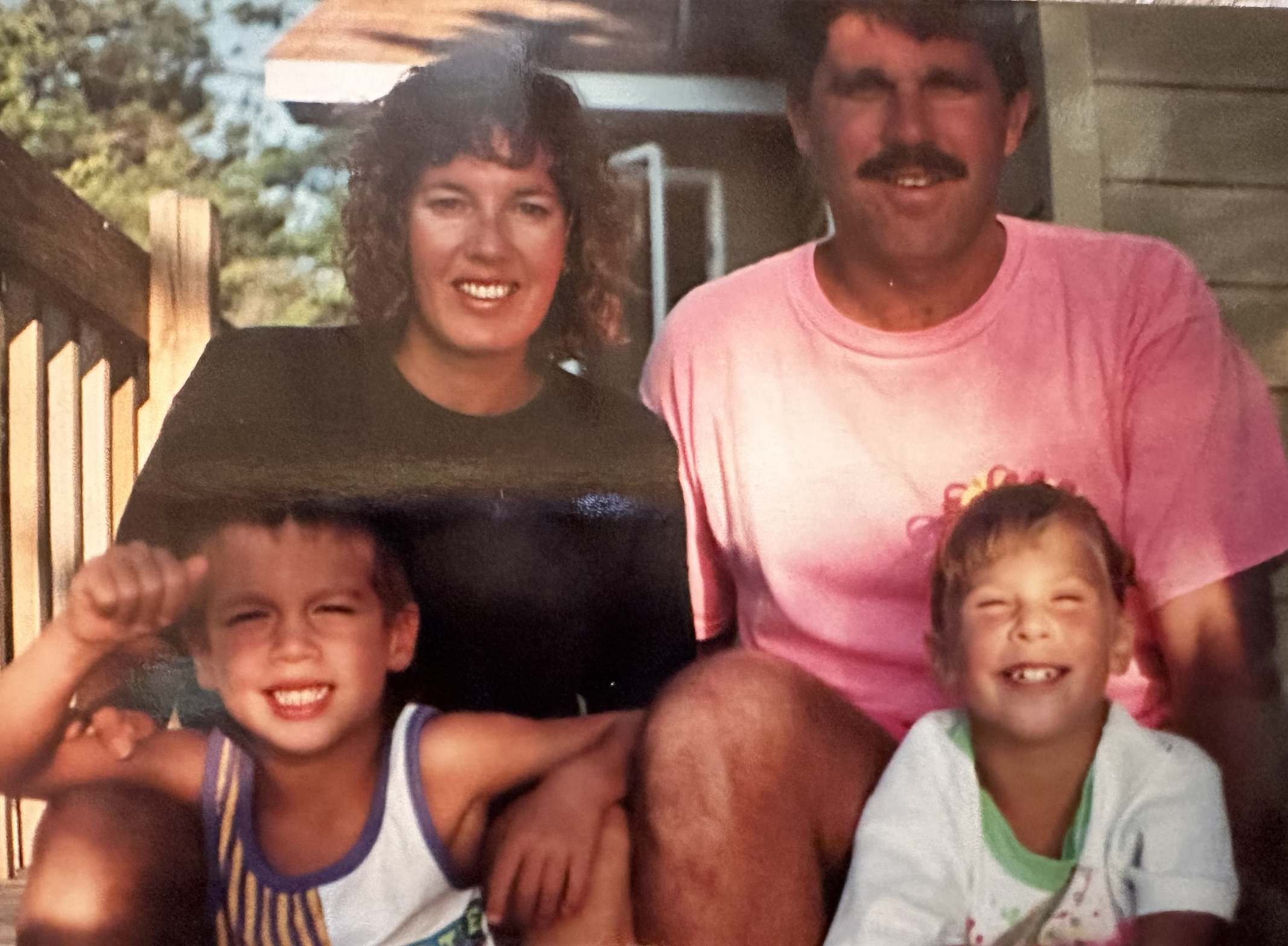 A family of four outdoors, with a woman and a man sitting together, and two young children in front. They are all smiling and appear happy.