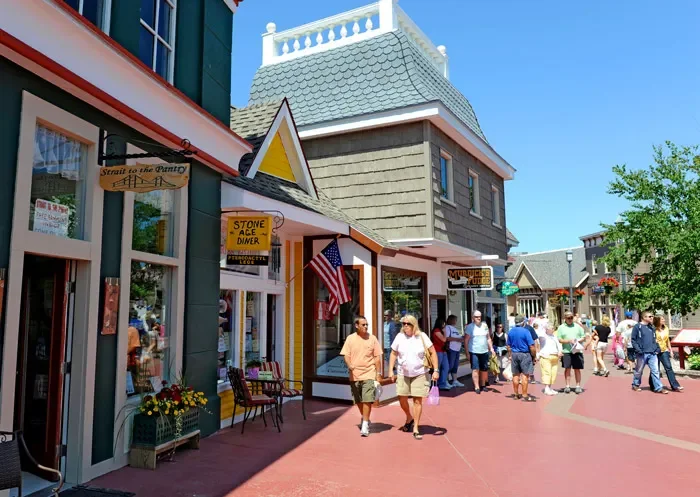  pedestrians walking along a storefront-lined street in a small town under clear blue skies, with colorful buildings, hanging flower baskets, and outdoor decor.