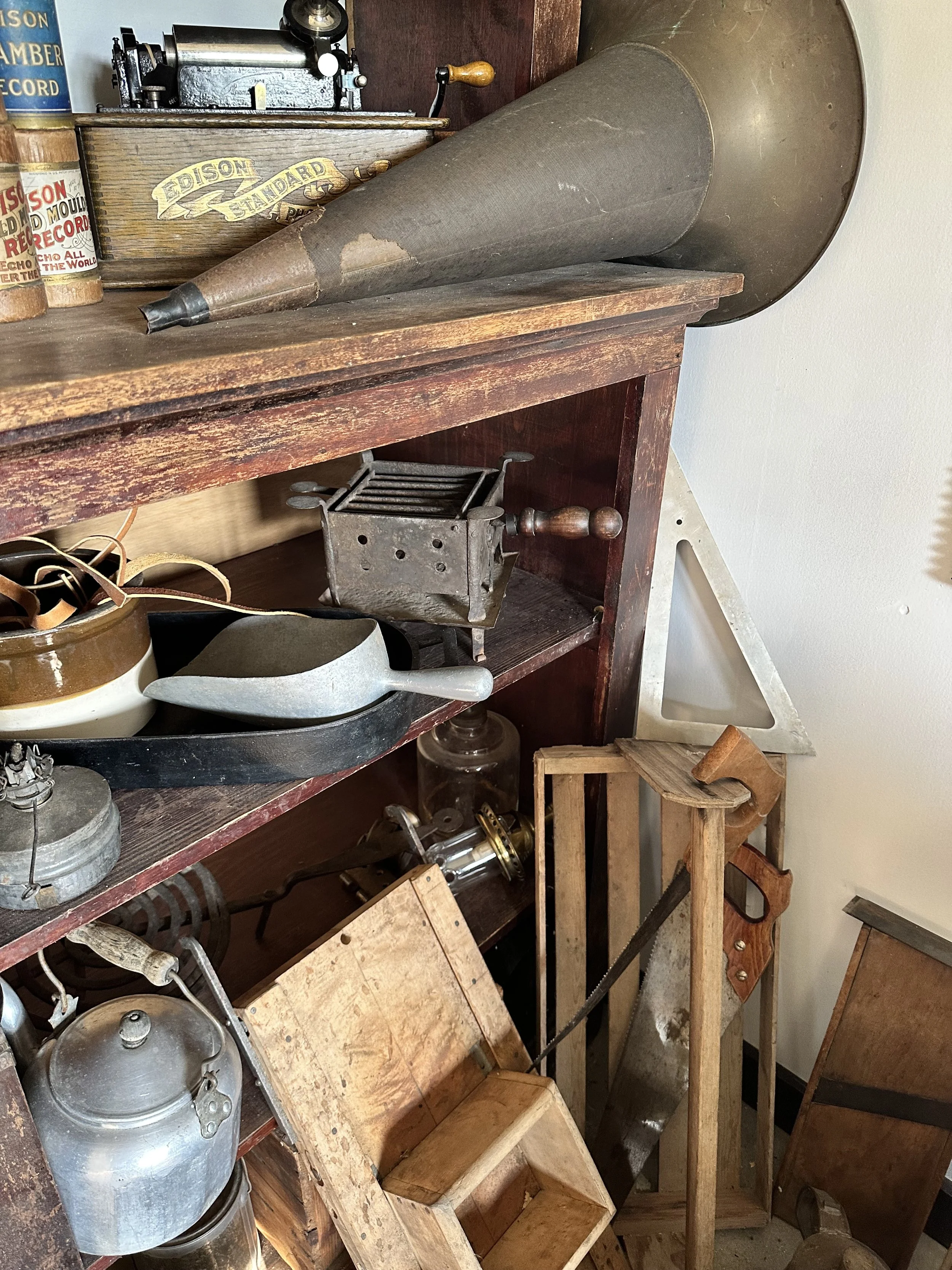 Old vintage objects on wooden shelves, including a gramophone, coffee grinder, metal kettle, wood and metal tools, and miscellaneous antique kitchen items.