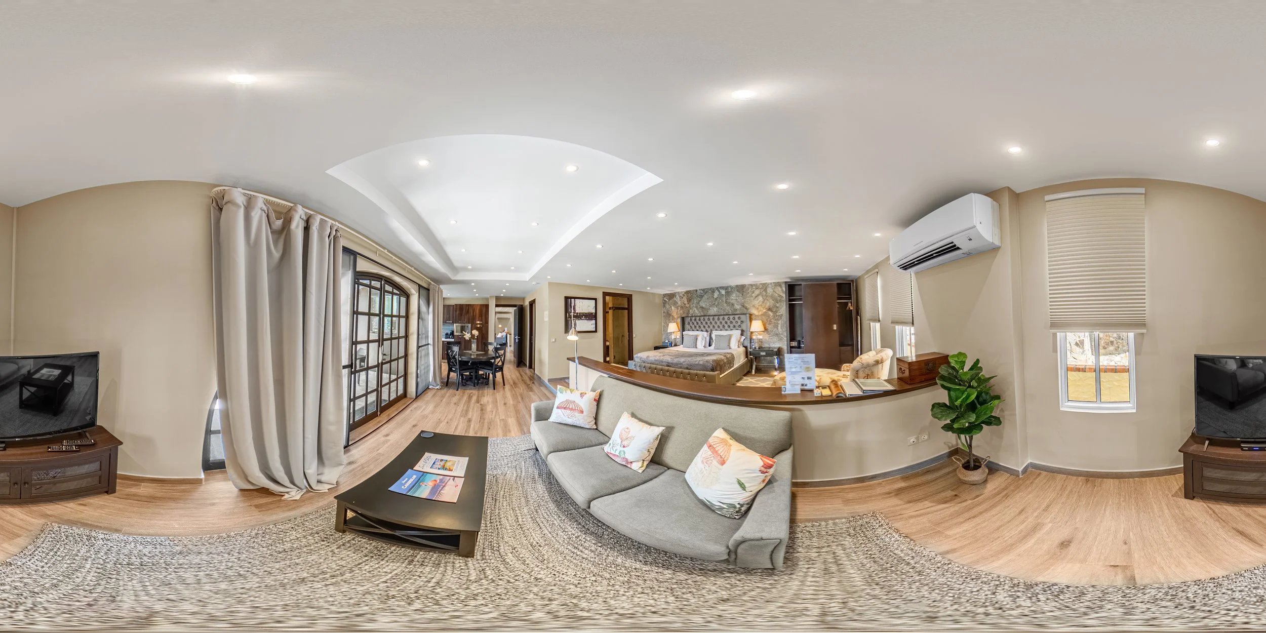 Spacious hotel suite with a living area and bedroom, featuring a sofa with decorative pillows, a coffee table with brochures, a TV stand with a television, large windows with curtains, and a sleeping area with a large bed, bedside lamps, and a stone accent wall.