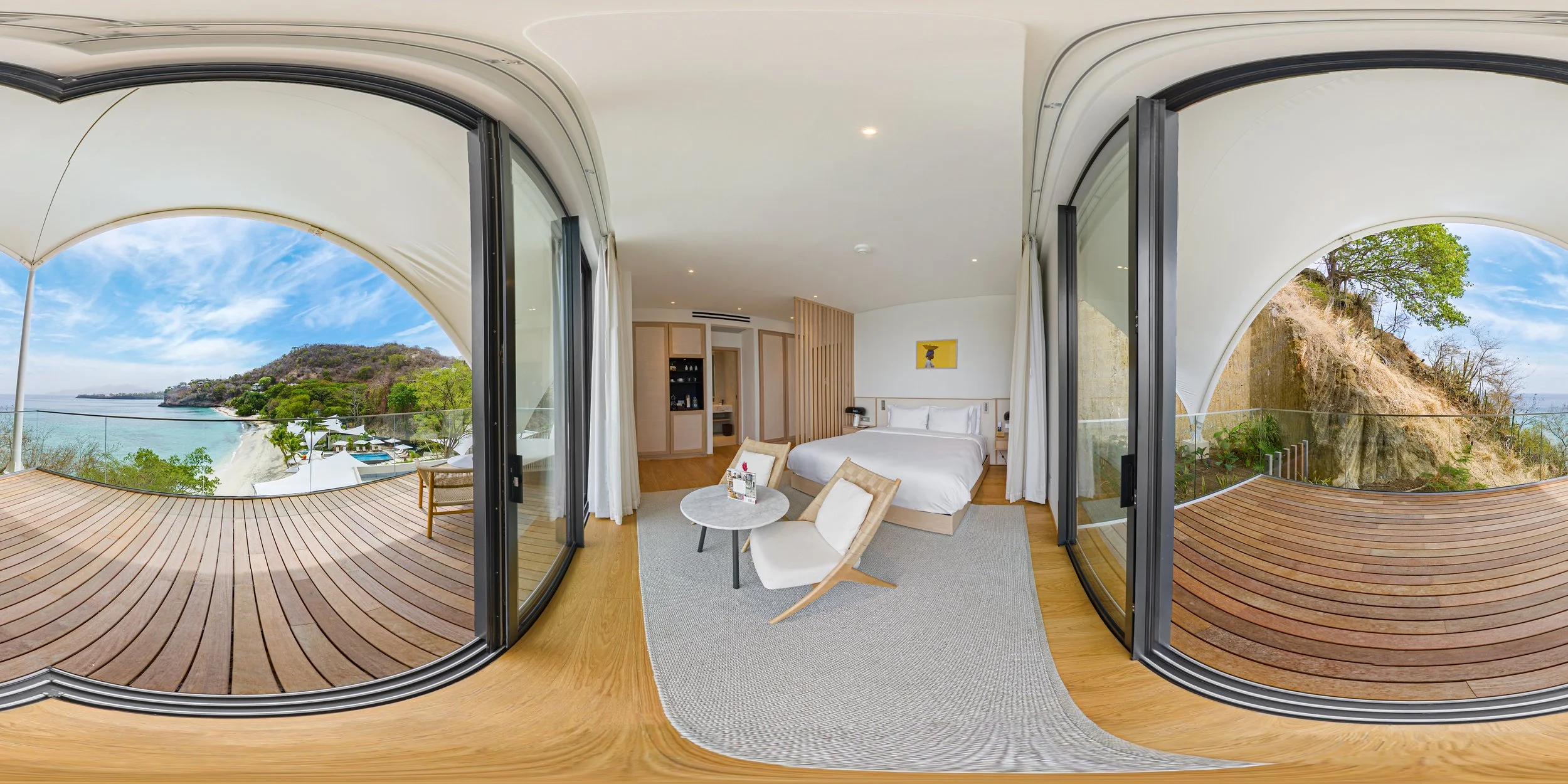 A hotel room with a large bed, a sitting area with two chairs and a small table, and a balcony with panoramic views of the ocean and hills.