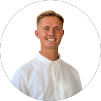 A young man with short blond hair, smiling, wearing a white collared shirt, standing against a plain white background.