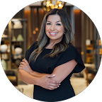 A woman with long dark hair smiling and standing with arms crossed in a modern indoor setting.