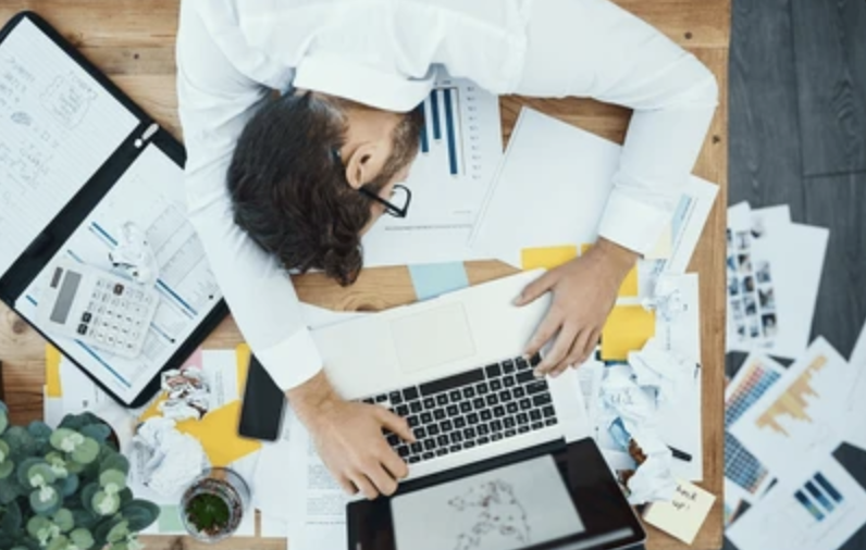 Man overwhelmed at cluttered desk with papers, tablet, and laptop.
