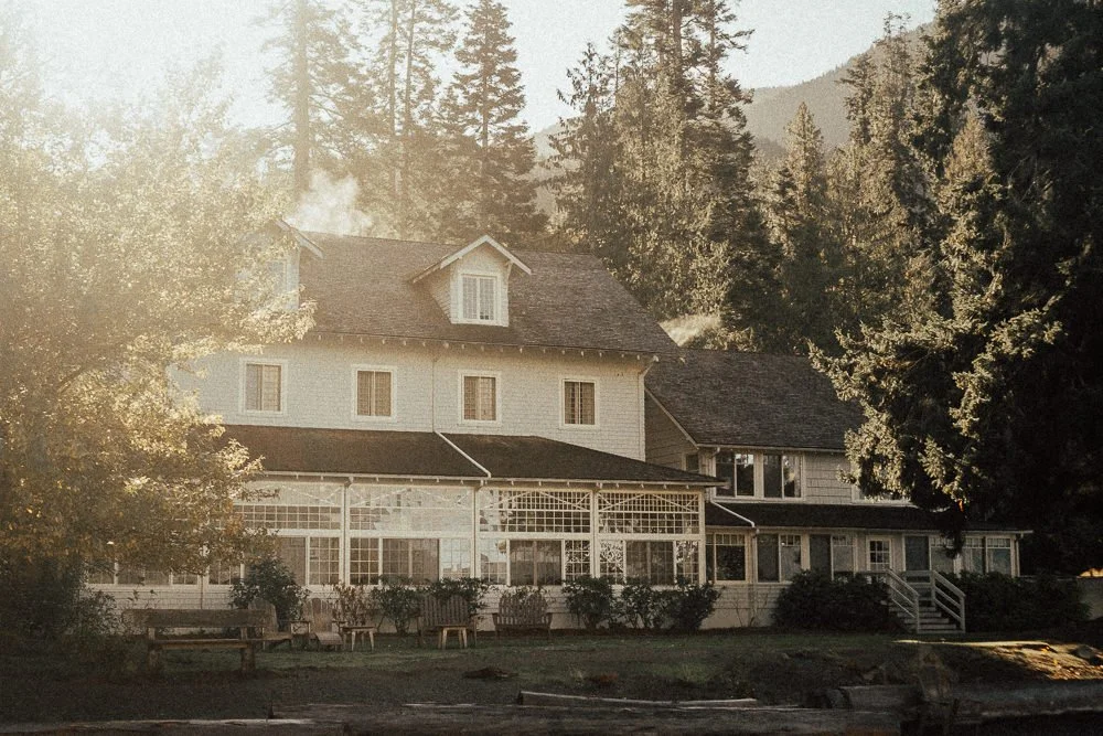 White building of Lake Crescent Lodge at Olympic National Park, Washington. A screened in porch set amongst trees in front of the mountains of Hurricane Ridge