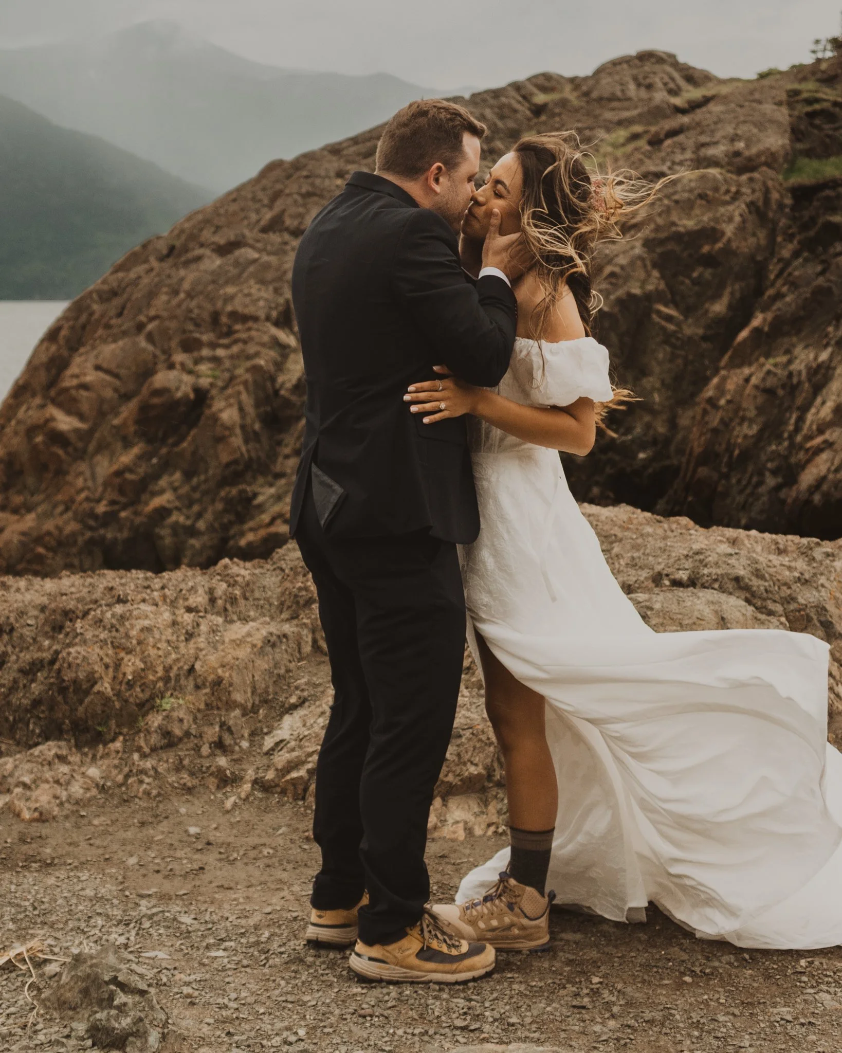 A man and woman are sharing an intimate moment outdoors, with the man gently holding the woman’s face as they touch noses, standing on rocky terrain with mountains in the background.