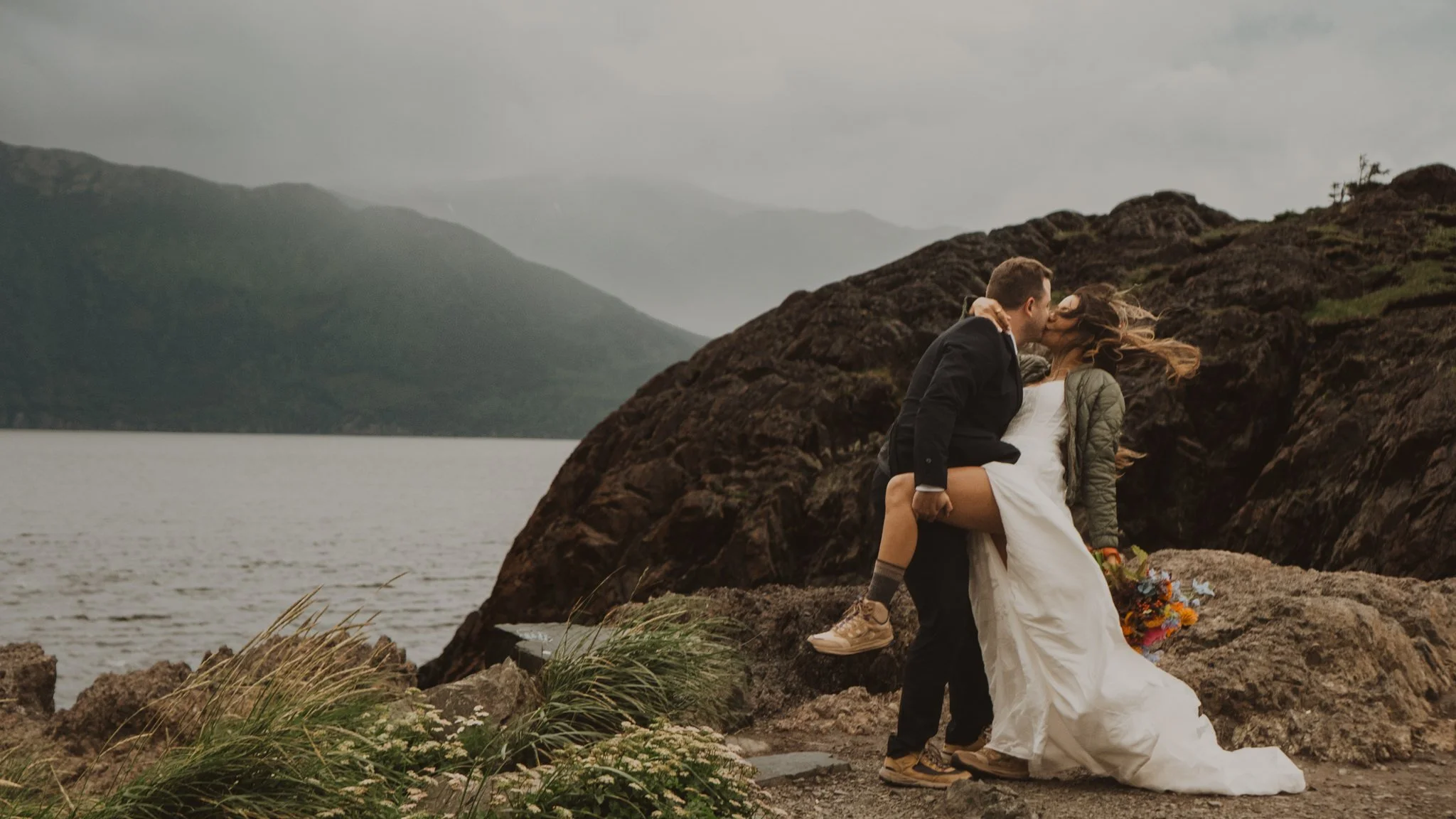 Groom lifting bride's leg while kissing her on a rocky Alaska shoreline, mountains and lake in the background