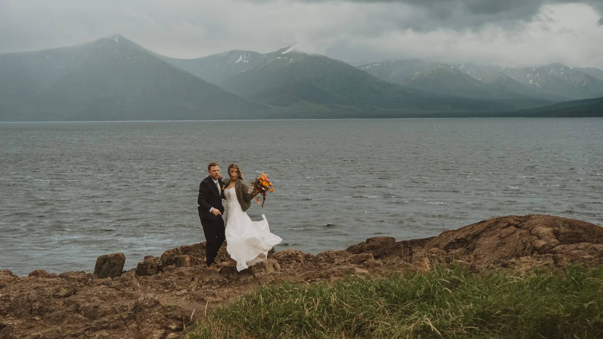 A bride and groom walk along rocky shore by a lake, with mountains and cloudy sky in the background. The bride holds a bouquet of flowers and wears a white wedding dress, while the groom is dressed in a suit.