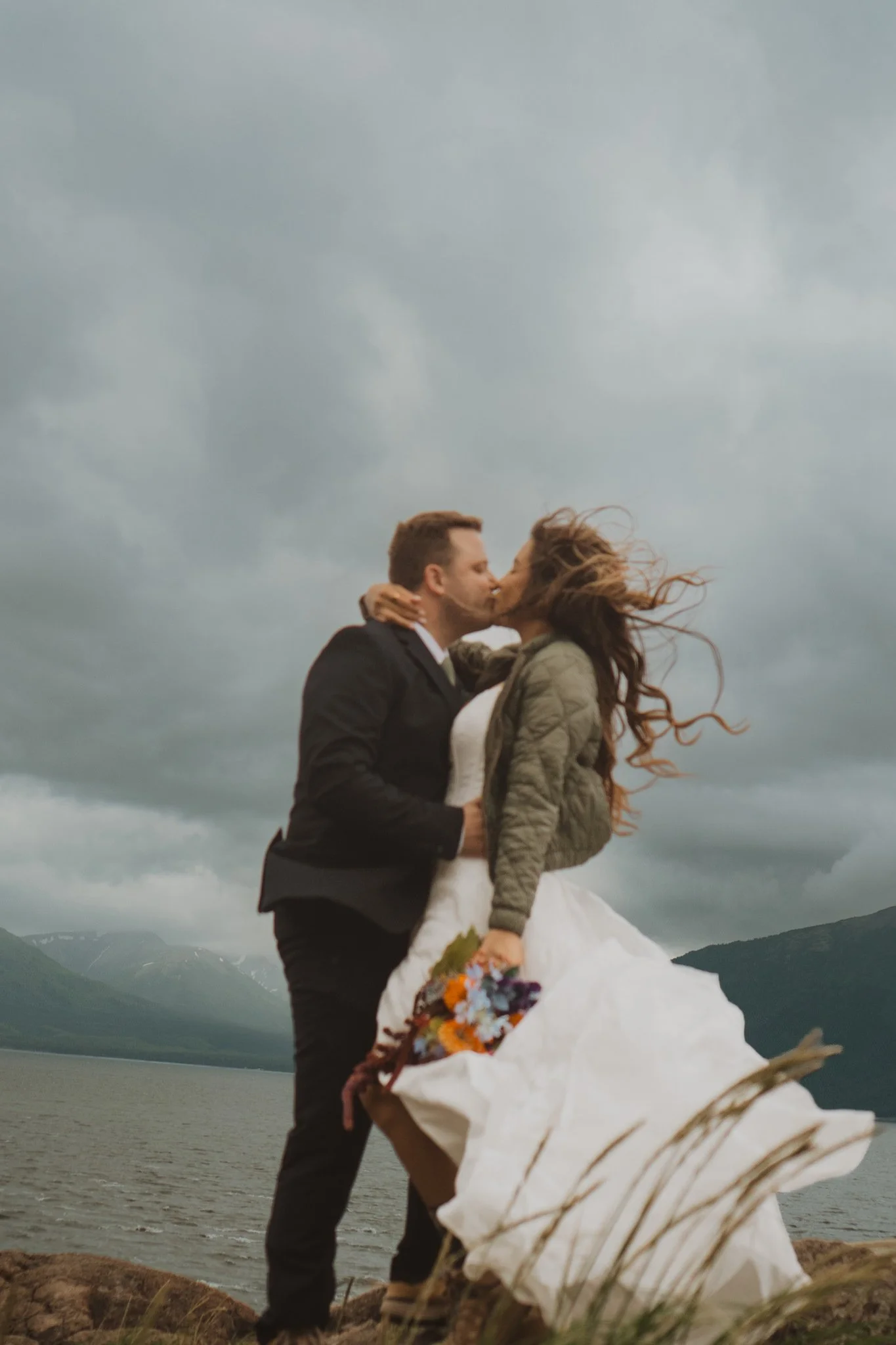 A man and woman kissing outdoors by a lake, with mountains and cloudy sky in the background. The woman holds a bouquet of flowers and wears a white dress and jacket. The man wears a black suit.