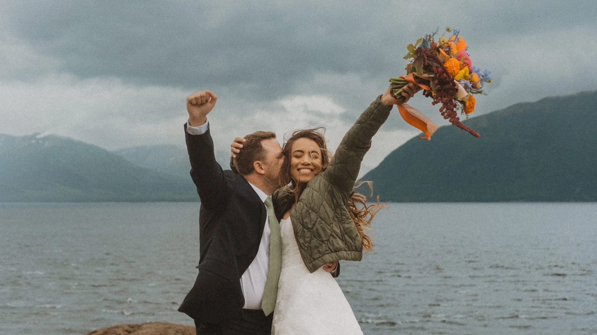 groom kissing bride with their fists in the air in front of mountains at BalugaPoint,Alaska