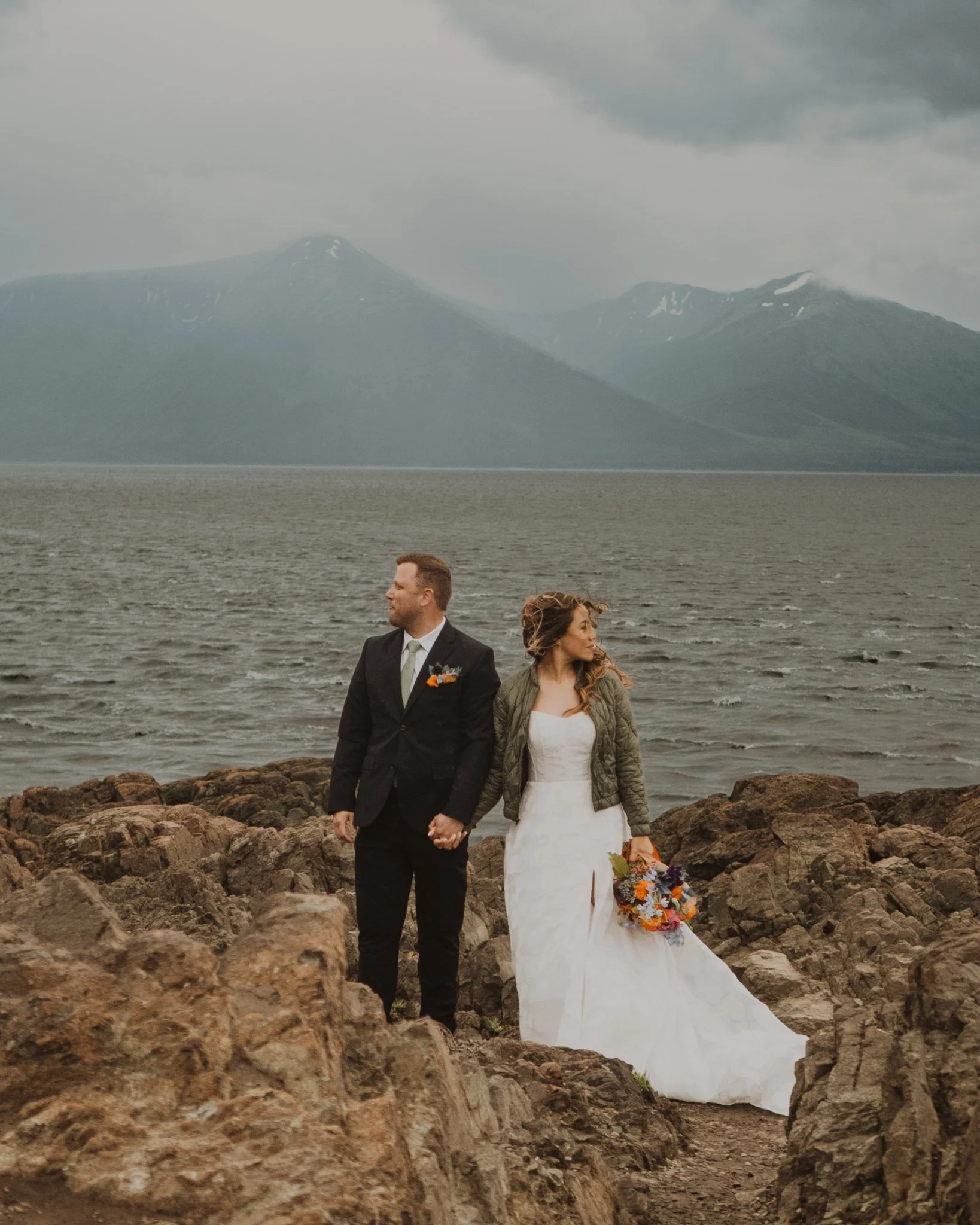 A newlywed couple holding hands, walking along a rocky shoreline near a body of water with mountains in the background under cloudy skies.