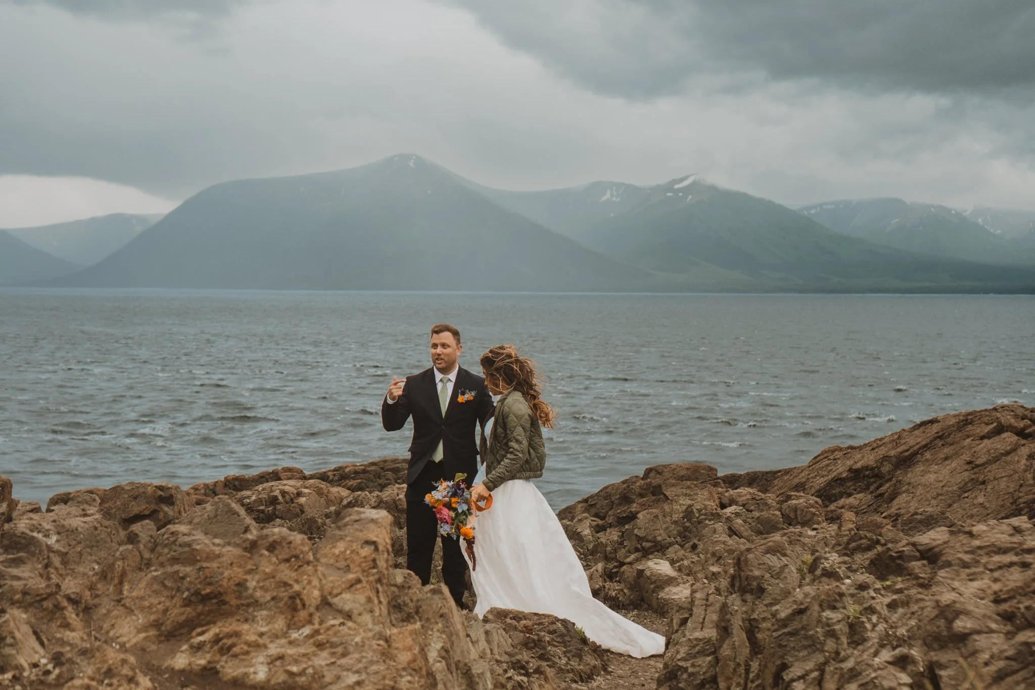 A couple dressed in wedding attire standing on rocky shoreline near water of Beluga Point, Alaska, with mountains and cloudy sky in the background.