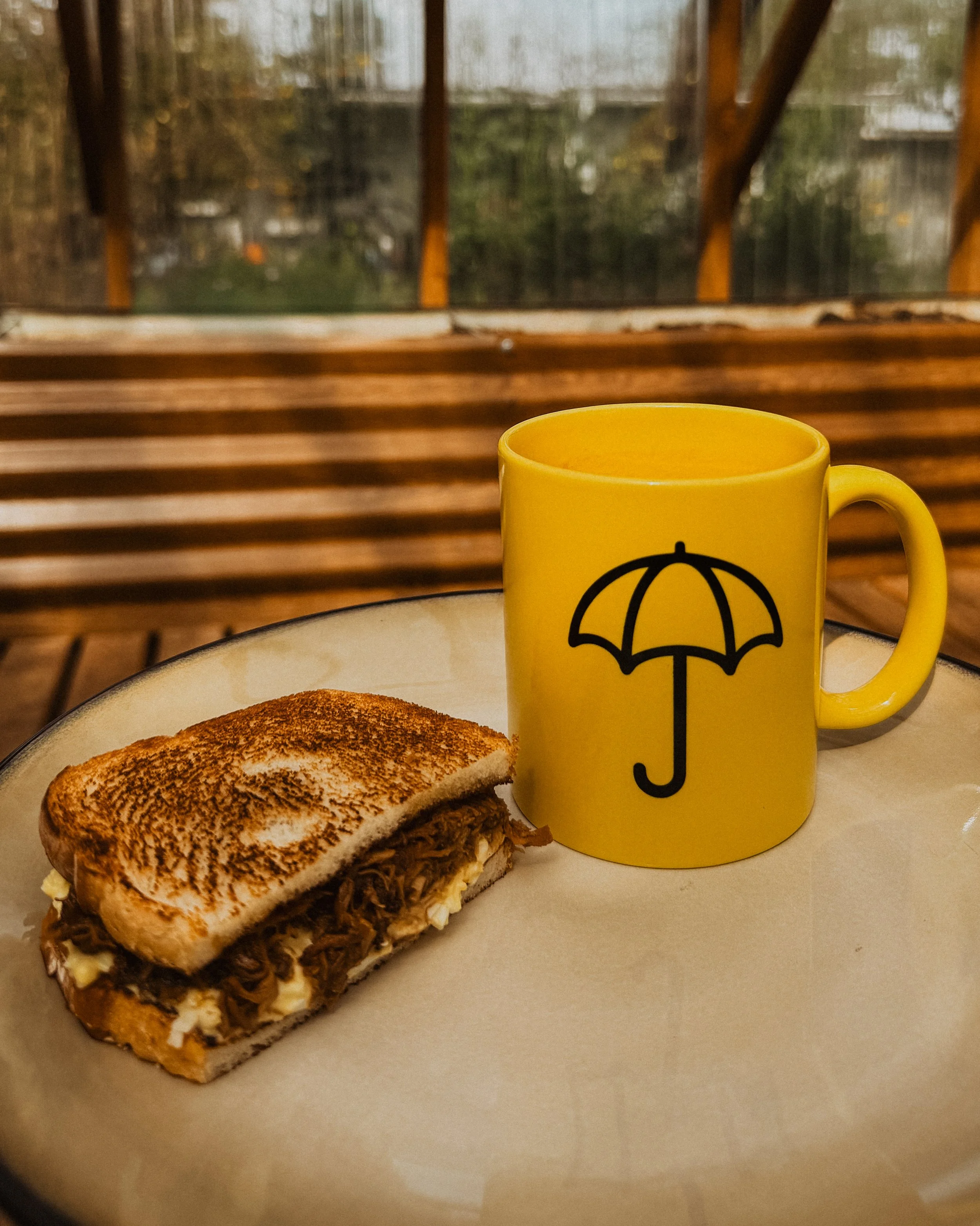 yellow coffee mug with black umbrella Fogtown logo plated with breakfast sandwhich inside a greenhouse