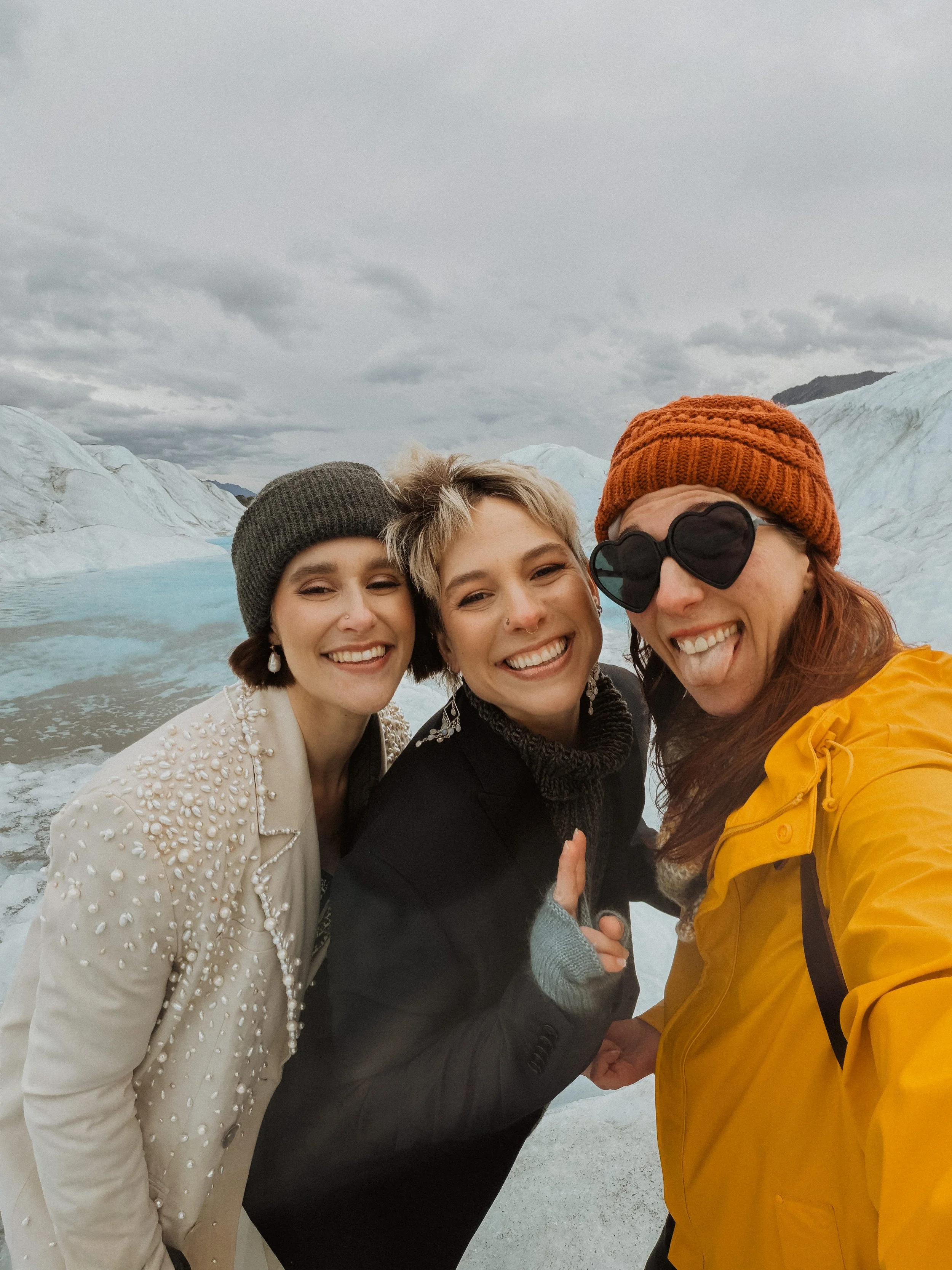 Four women smiling and posing for a selfie outdoors near a glacier with ice mountains in the background, dressed in winter clothing.