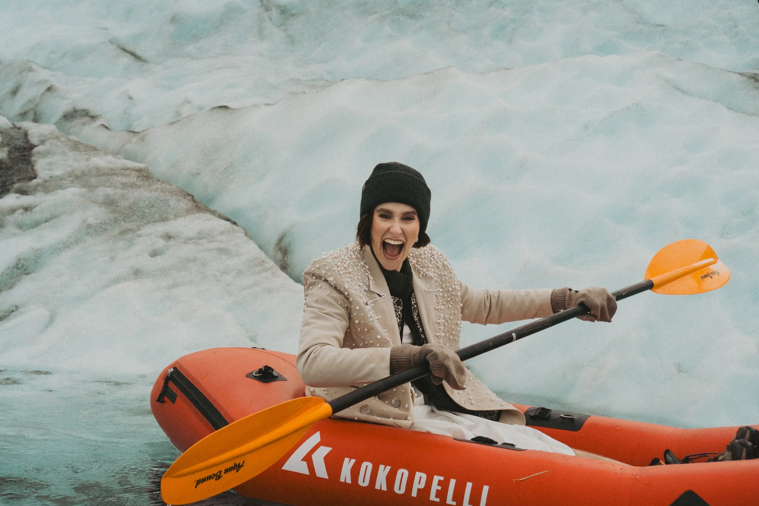 A woman in a beige coat with pearl embellishments, a black beanie, and gloves, is smiling and paddling a kayak on icy water of Knik glacier. The kayak is orange with black and white markings, and the woman is holding an orange paddle.