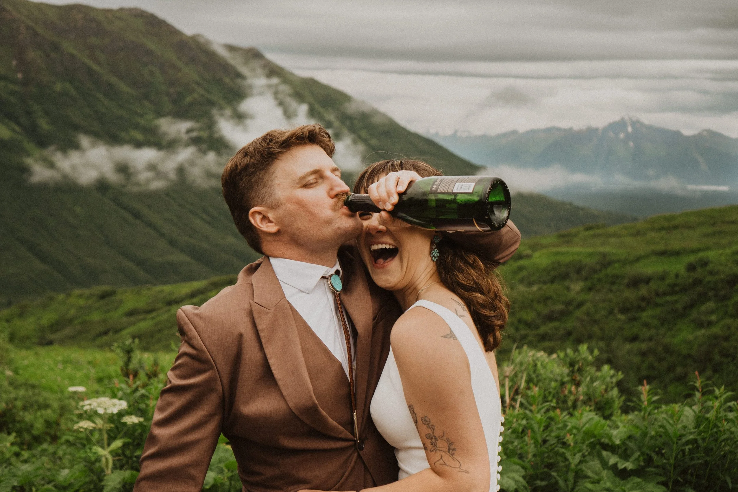 Couple in wedding attire drinking champagne together on a lush green mountainside after eloping in Alaska