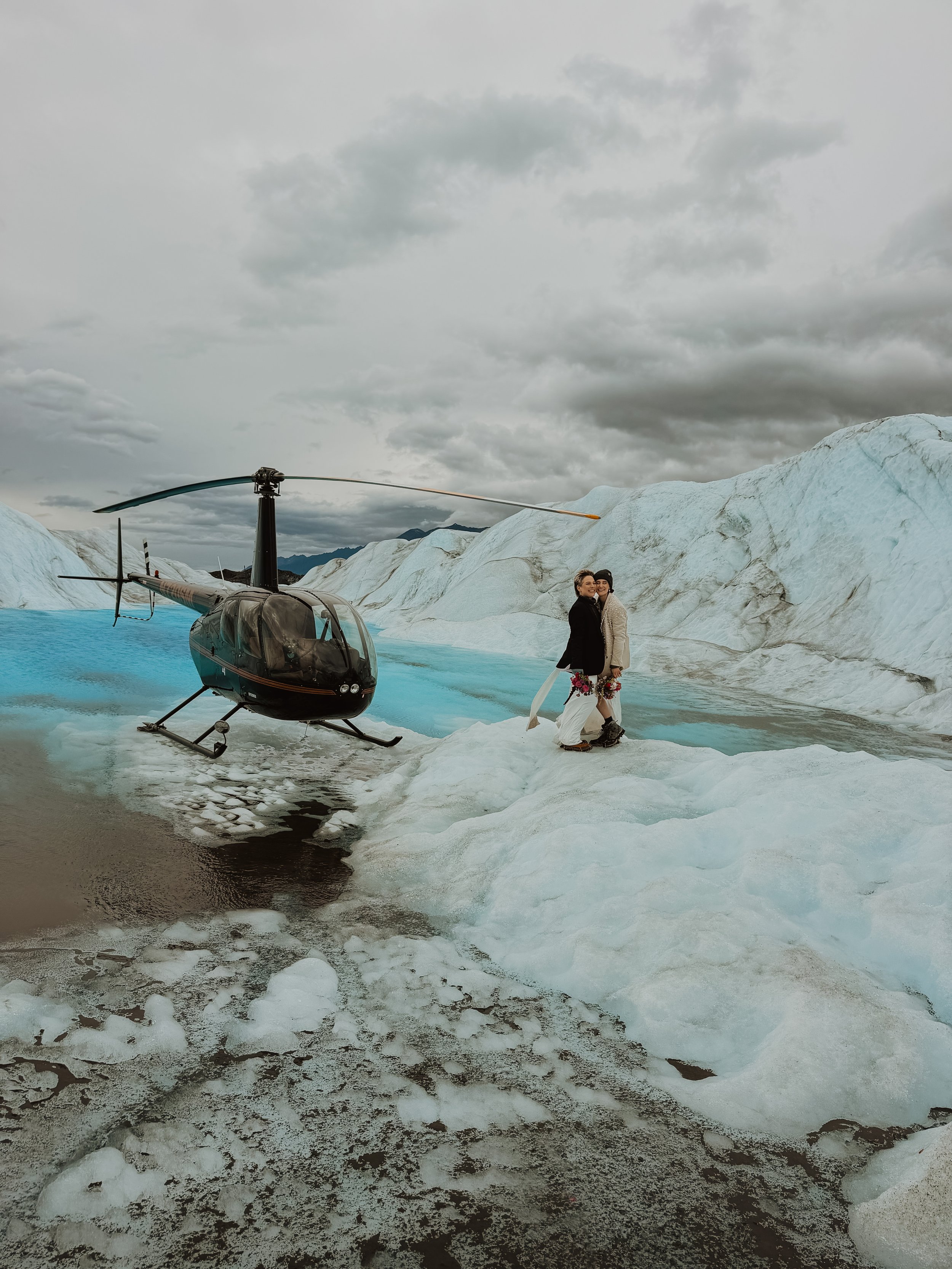 Two women in winter clothing standing on icy terrain near a black helicopter with a glacier and cloudy sky in the background.