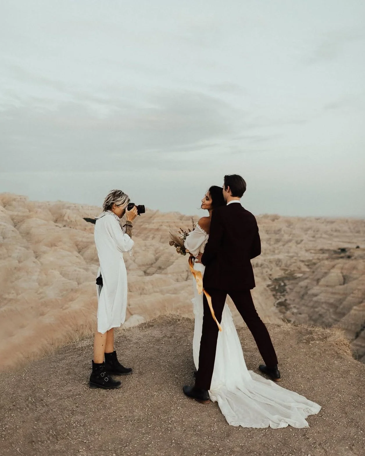 photographer Paige Kleckner taking a photo of an elopement couple of a man in a marooon suit standing behind bride wearing long white dress his hands on her hips posing on a bluff in Badlands National Park