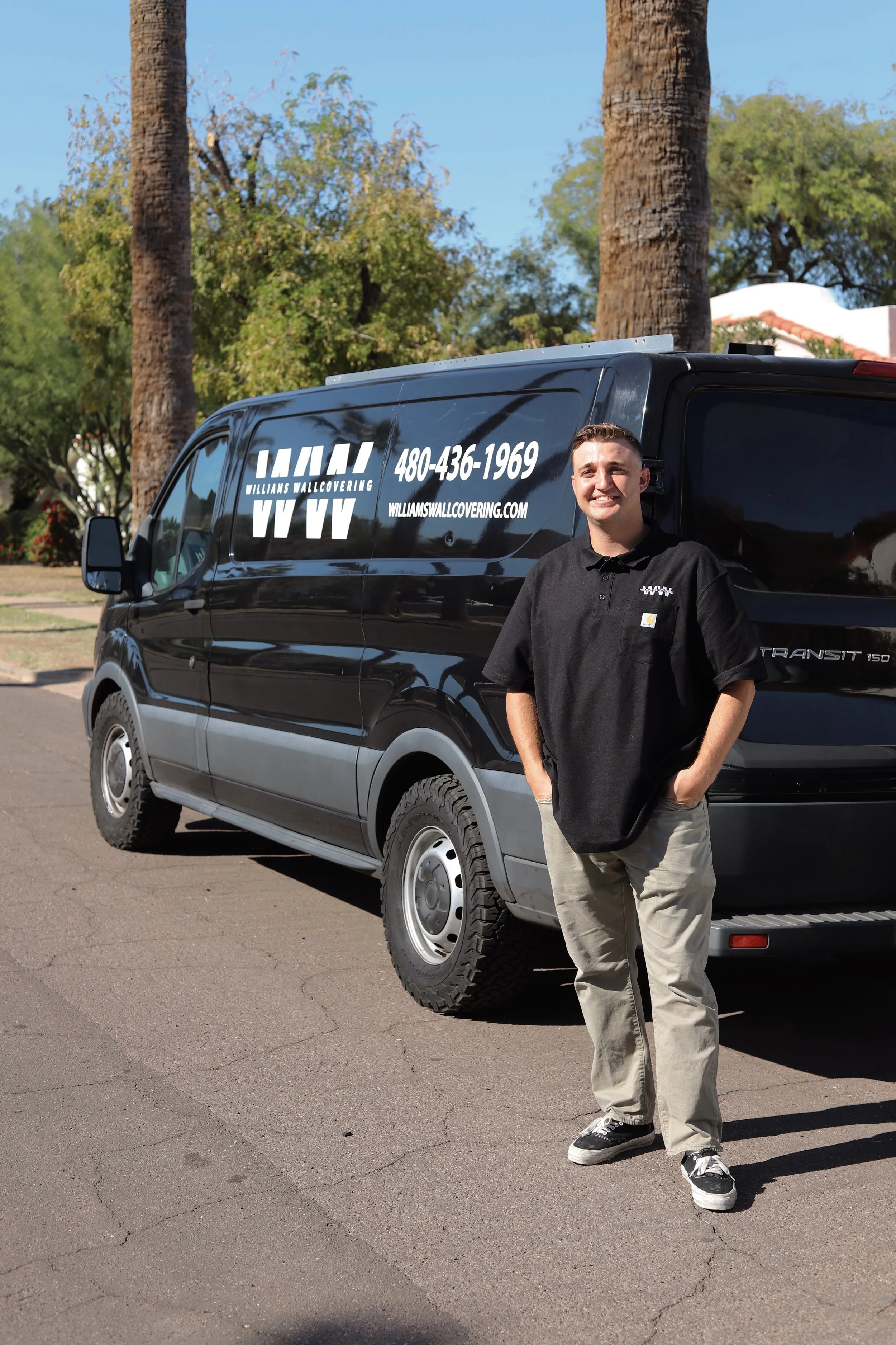 A man stands next to a black van with signage for Williams Wall Covering, containing a logo, phone number, and website, on a sunny day with palm trees in the background.