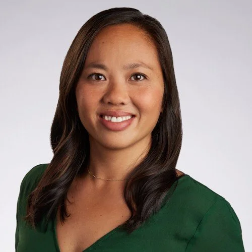 Portrait of a smiling woman with dark hair wearing a green top against a plain light gray background.