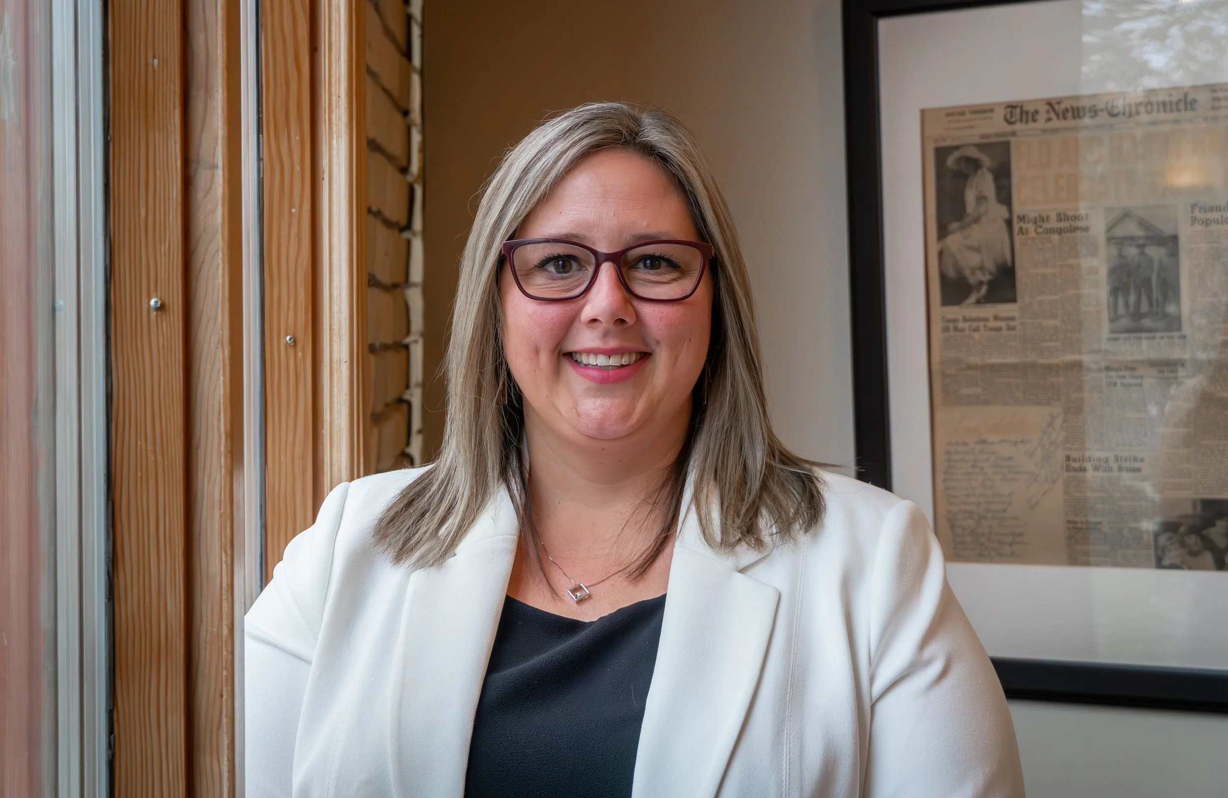 Professional woman with glasses and shoulder-length hair smiling at the camera, wearing a white blazer and black top, seated indoors with a framed newspaper on the wall behind her.