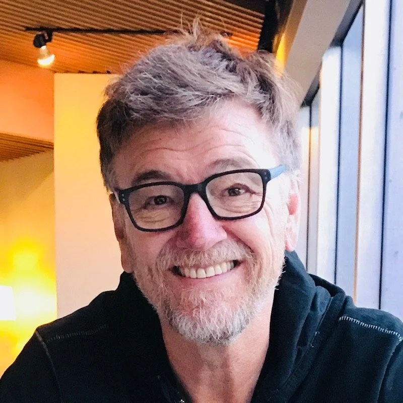 Smiling man with glasses, gray hair, and beard, sitting near large windows in a modern, warmly lit interior.
