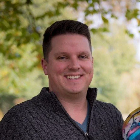 A smiling man with short dark hair wearing a black zip-up sweater outdoors with green and yellow fall foliage in the background.