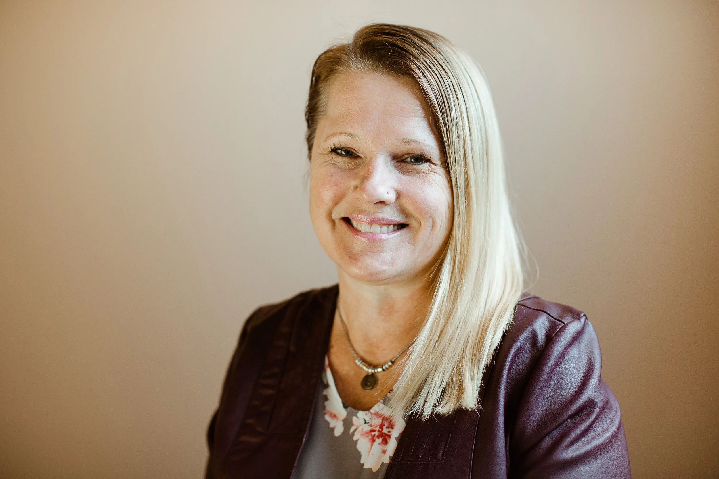 A woman with blonde hair smiling in front of a beige background, wearing a maroon leather jacket and a floral top.