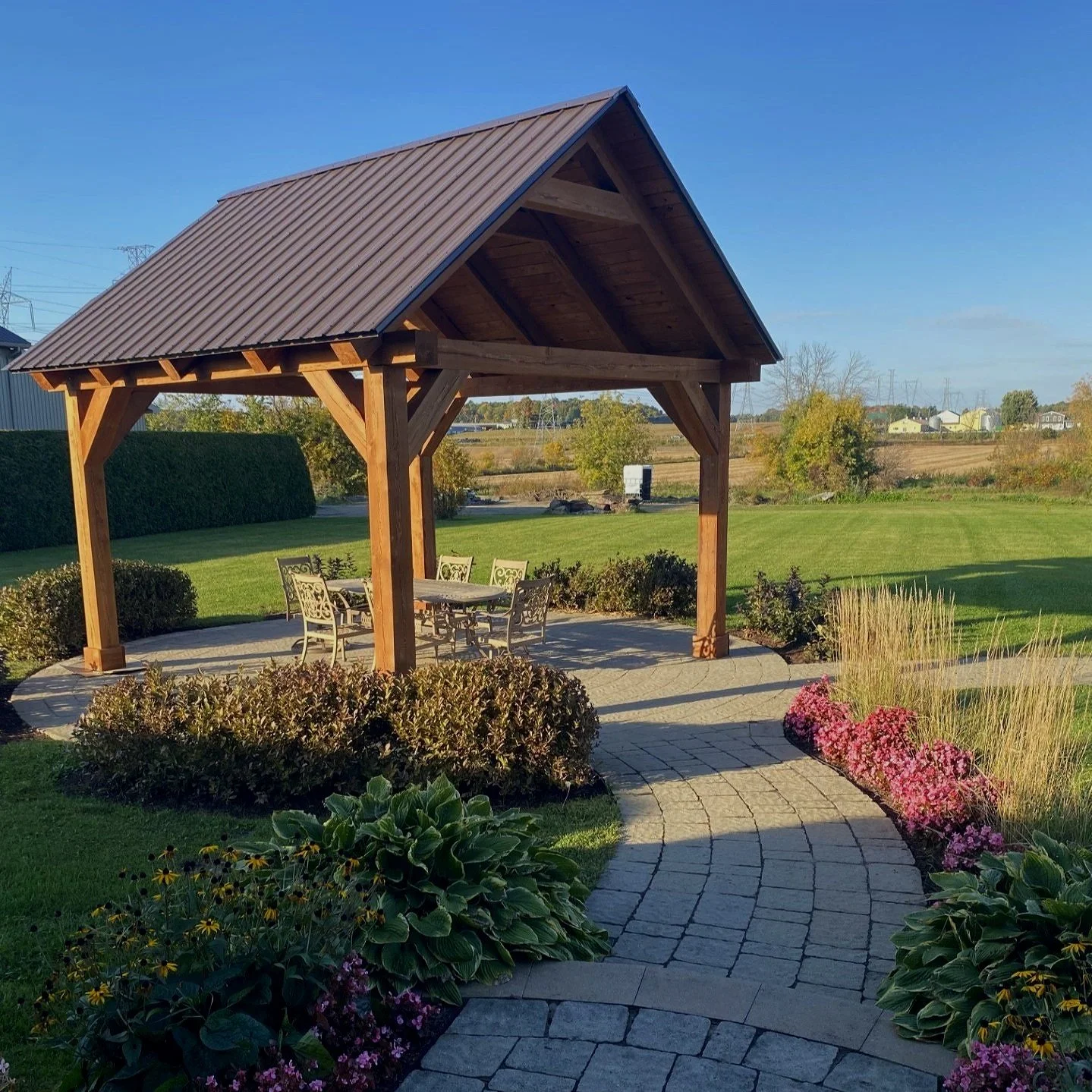 Une pergola en bois avec une toiture en métal, entourée d'un jardin bien entretenu avec un chemin en pavés, des arbustes, des fleurs colorées et une pelouse verte, sous un ciel bleu clair.