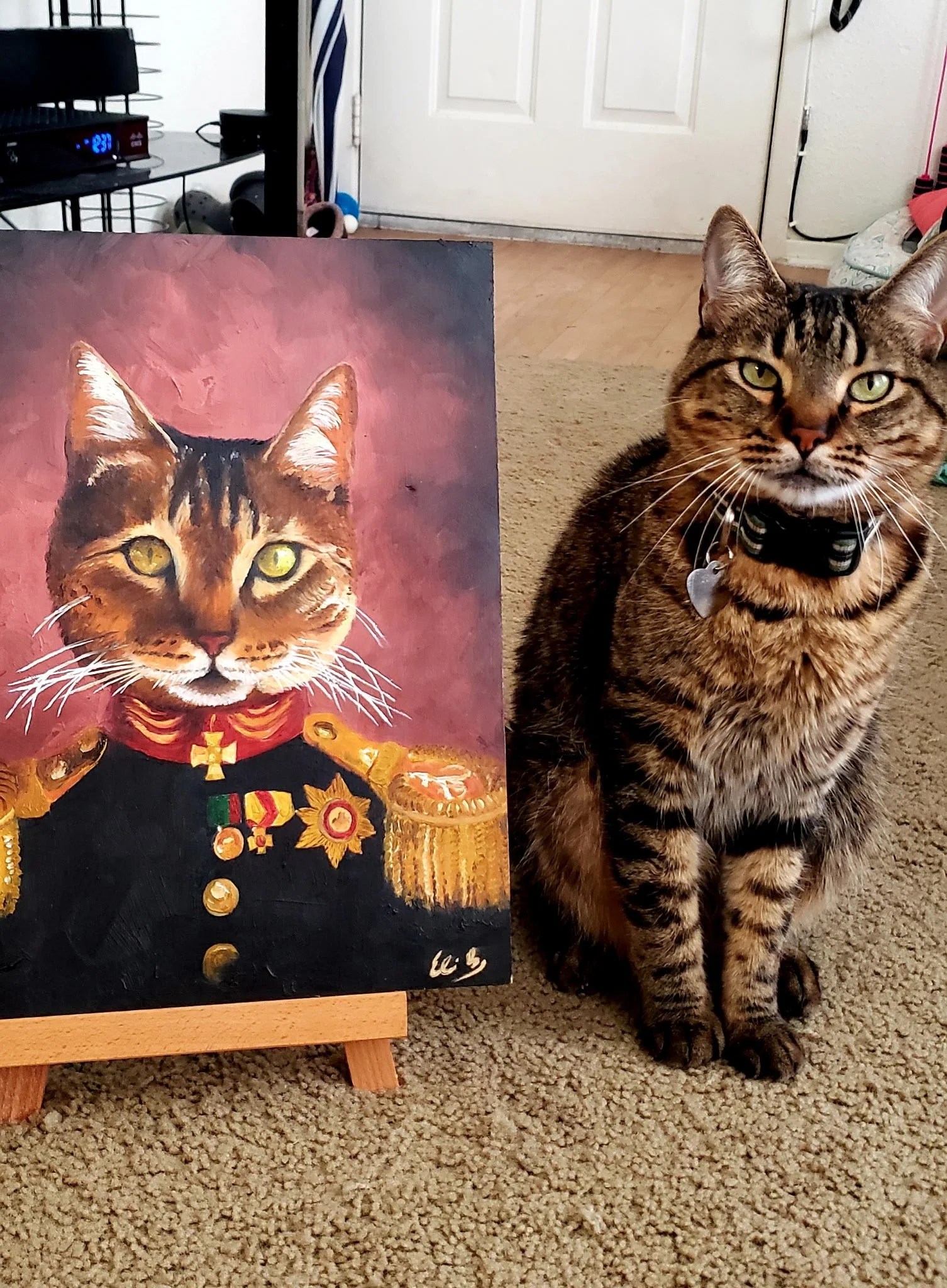 A tabby cat with green eyes wearing a black collar with a heart-shaped tag sitting next to a painting of a cat dressed as royalty. The painting depicts a cat with a similar appearance, wearing a red and gold royal uniform with medals and gold trim.