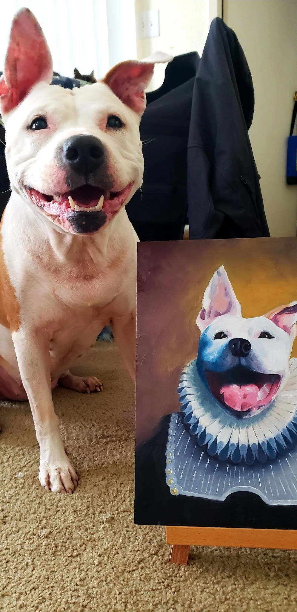 A happy white dog with a black nose sitting on a beige carpet next to a painting of a dog with a ruffled collar and pearls. There is a black backpack with clothing hanging on a chair in the background.